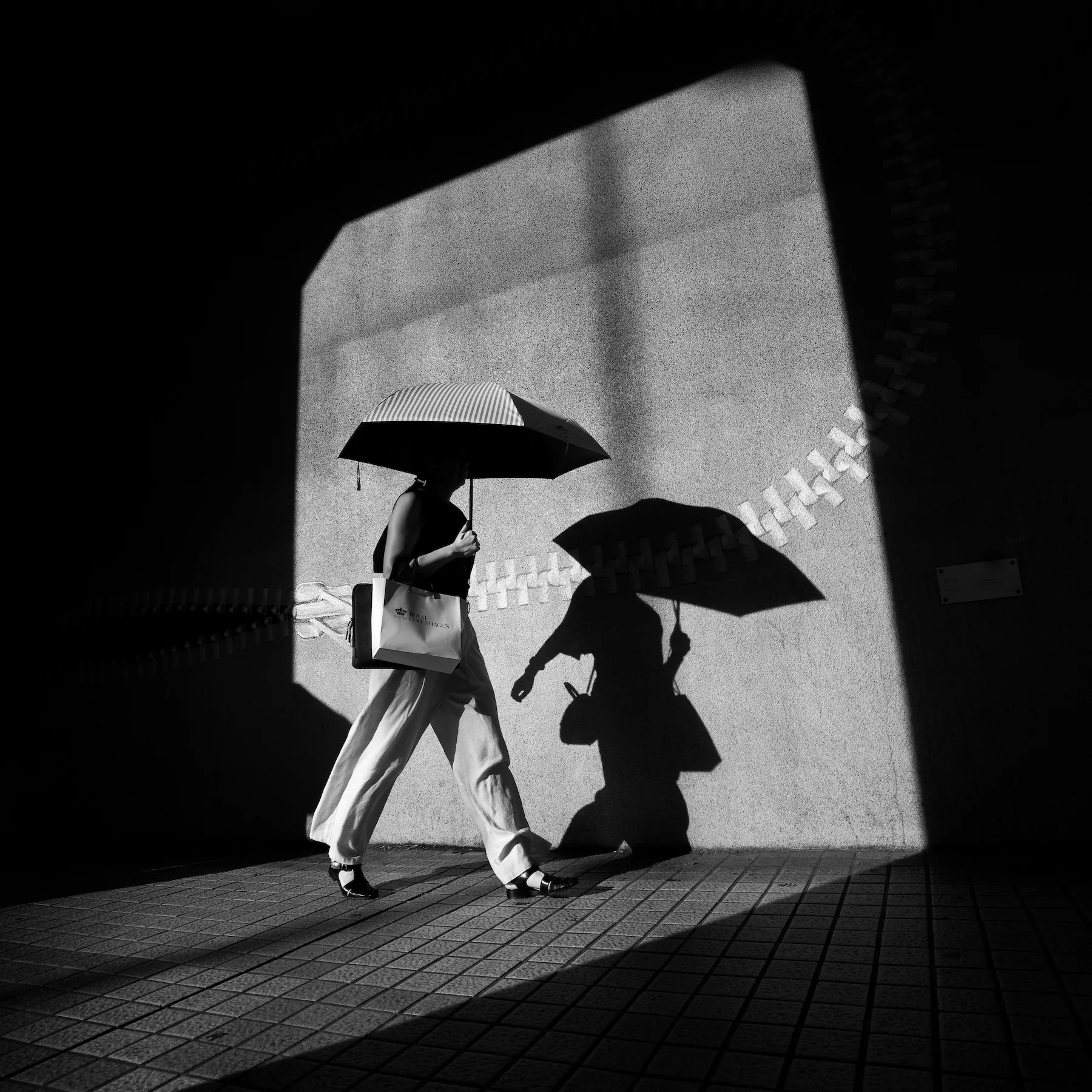 A woman walking on a tiled street holding an umbrella, with her shadow cast on a textured wall, framed by a dark doorway.