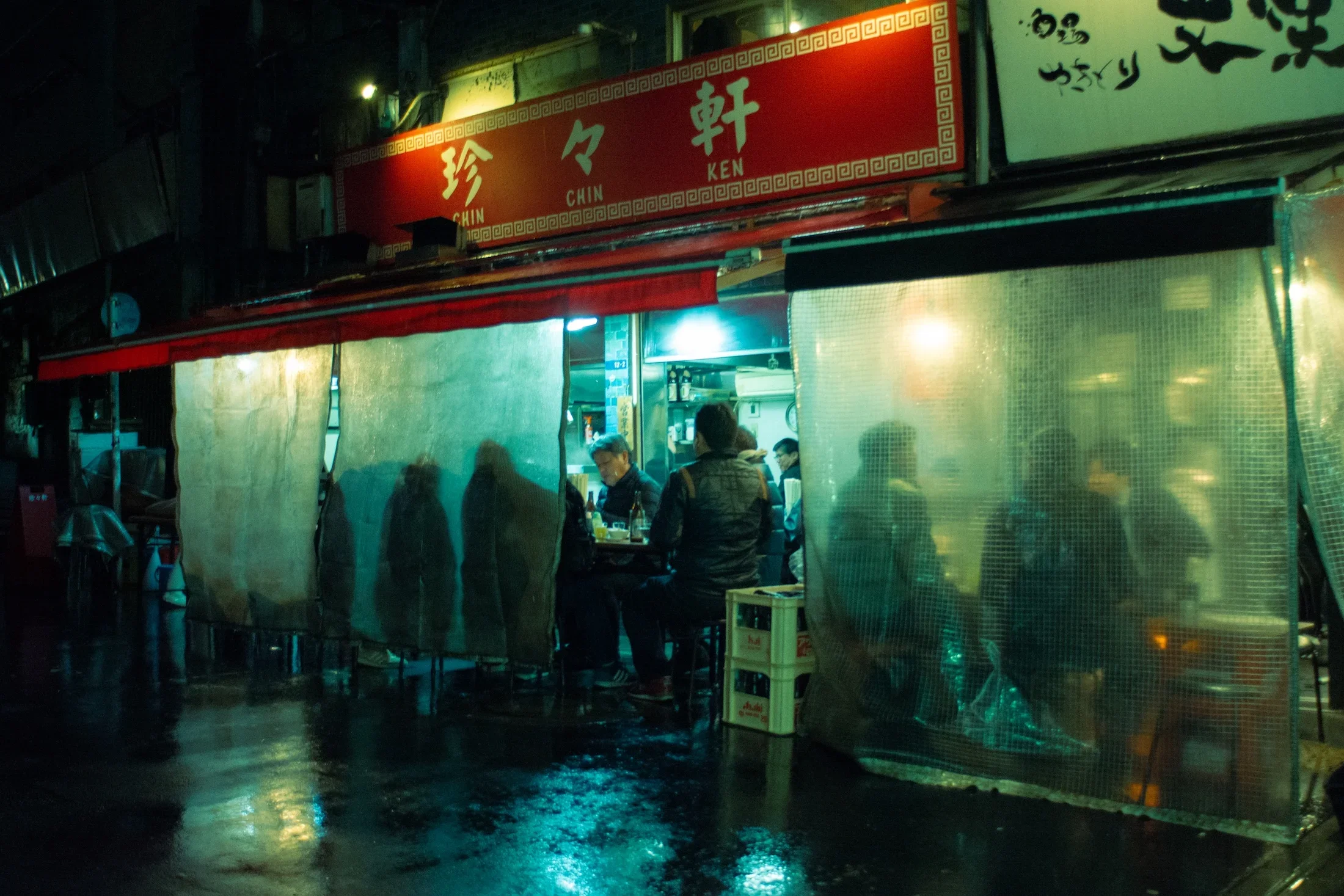 Nighttime street scene showing a small Asian restaurant with a red sign featuring Chinese characters and the words 'Chin Ken.' Inside, several people are seated and dining, with some visible through the open restaurant entrance.