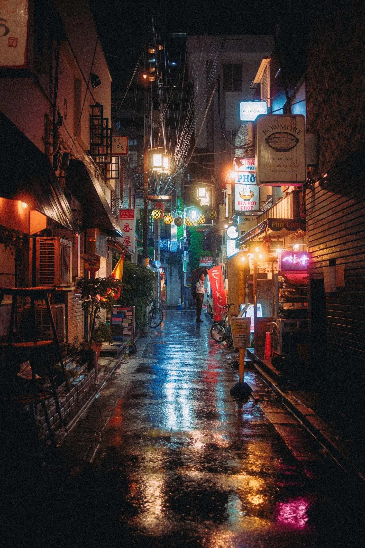 A narrow street at night with wet pavement reflecting colorful neon lights and signs. Small storefronts and hanging lanterns line the street, with a person holding an umbrella walking in the distance.