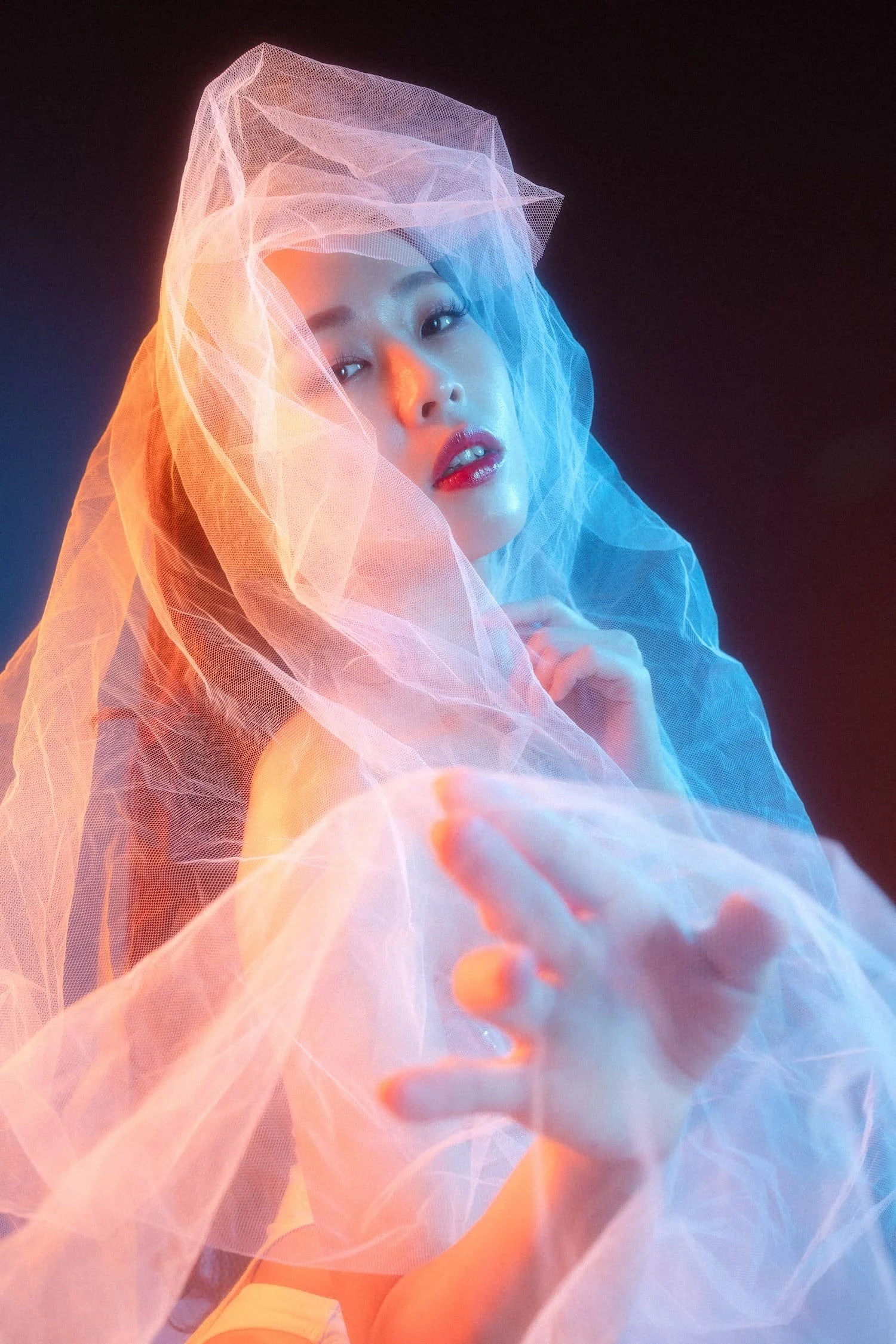 A woman with makeup and red lipstick is partially covered with sheer, colored fabric, looking at the camera with her hand extended forward, against a black background.