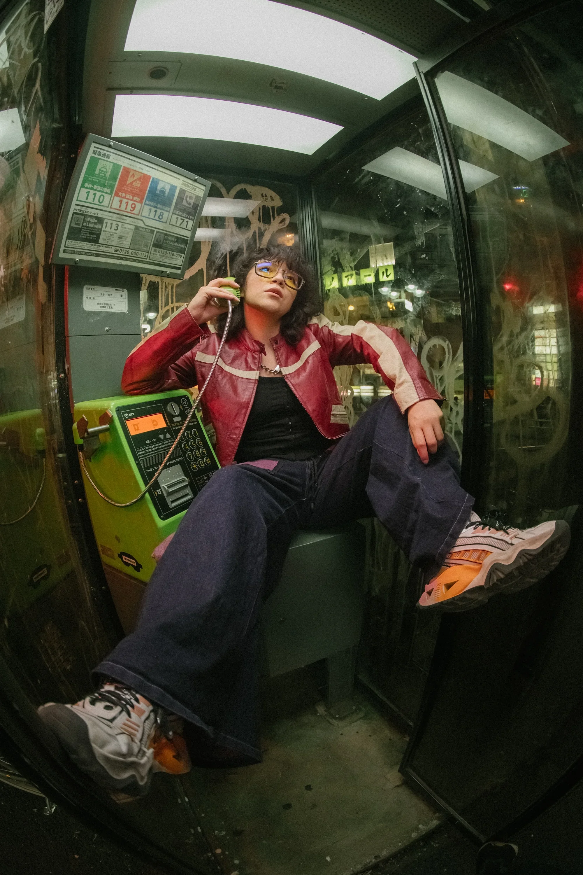 A young woman with glasses, curly hair, and a red and beige jacket, sitting inside a small glass booth at night, talking on a green payphone. She is wearing dark jeans and sneakers, with one foot resting inside the booth. The booth has a digital display and a sign with phone numbers on a monitor behind her. The surrounding environment is dark with some lights visible outside.