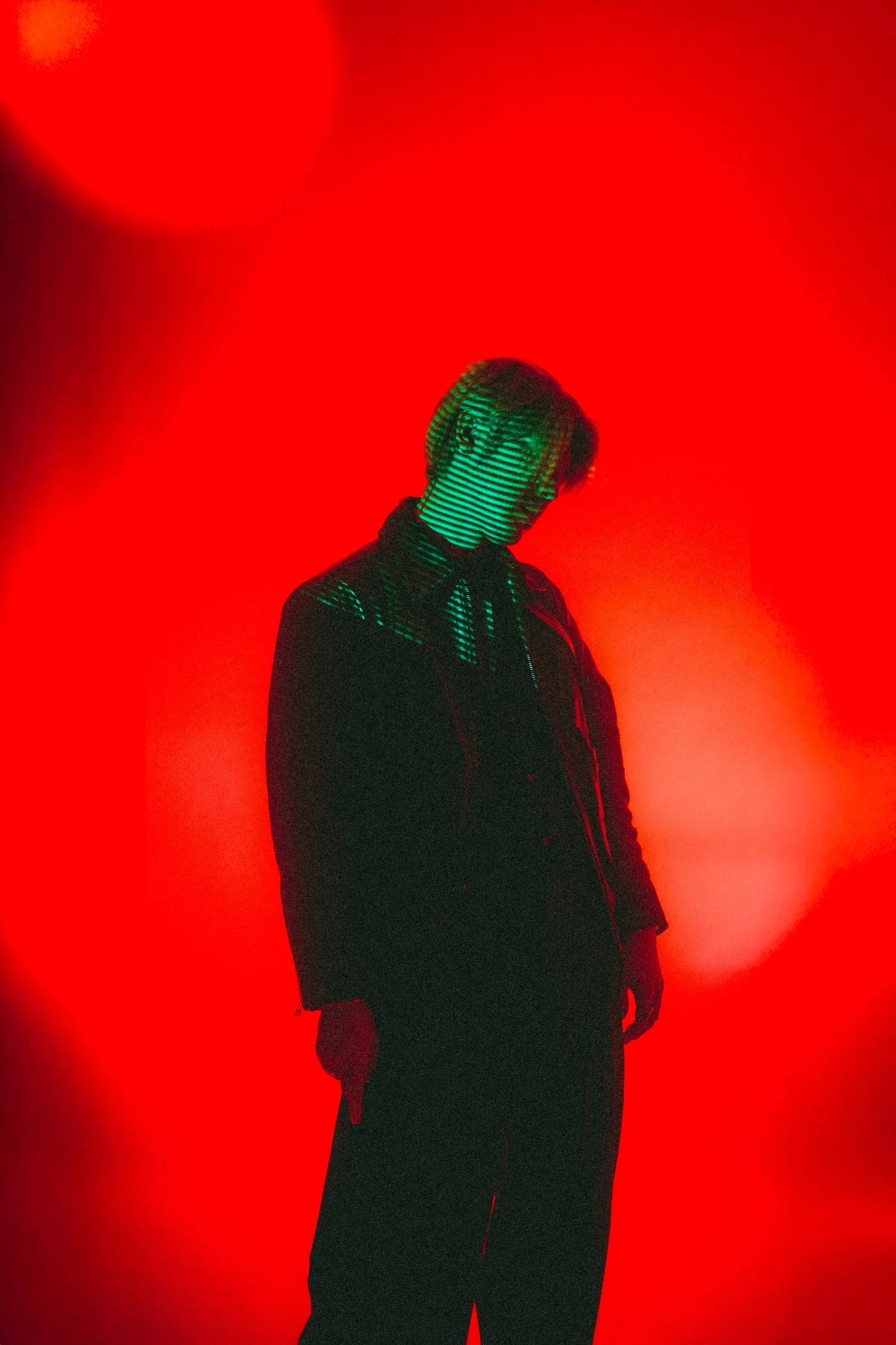 A person with short hair is standing against a vibrant red background, with a green laser light projected onto their face and upper body, creating a striped pattern.
