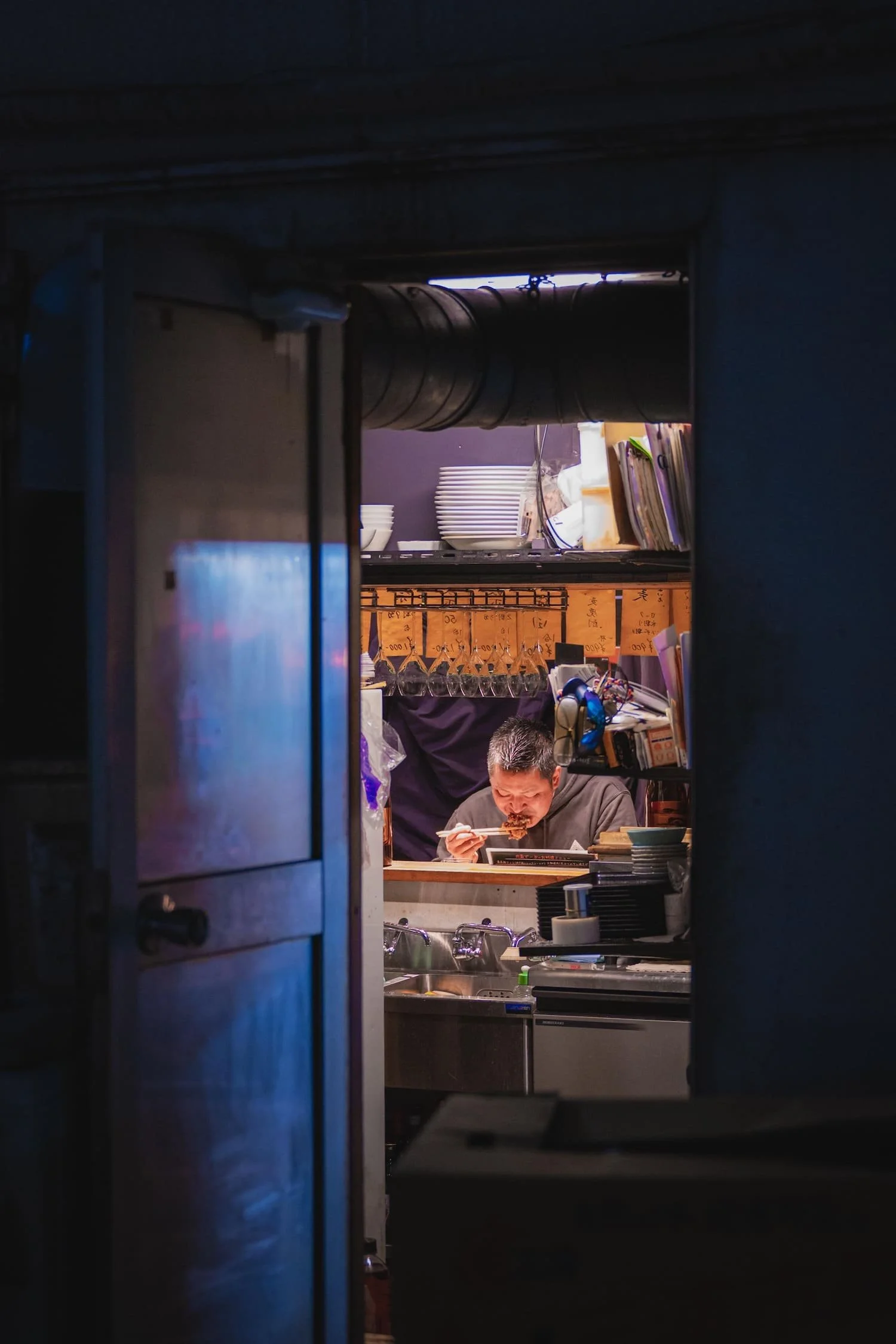 A man eating ramen in a restaurant kitchen, viewed through a doorway.