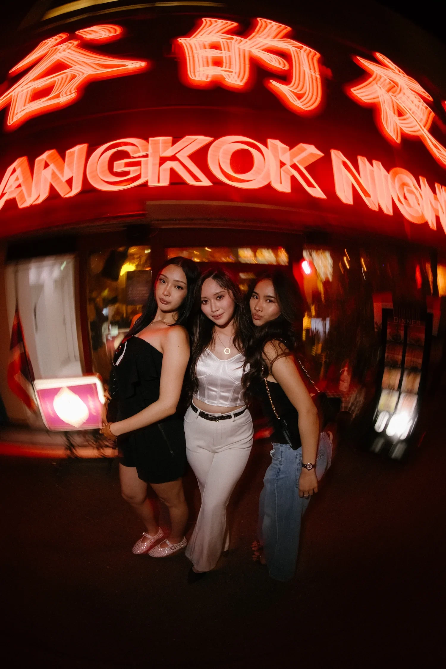 Three young women standing in front of a neon sign that reads 'Hong Kong N' with additional characters obscured, outside a restaurant at night.