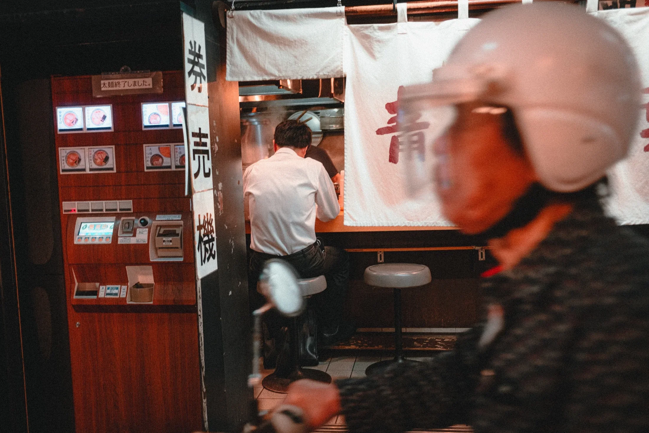 A person wearing a white shirt is working behind a counter in a Japanese restaurant or food stand. There are menus with images of food on the wall to the left, and a white curtain with Japanese characters is hanging in front of the workspace. The scene appears to be indoors with warm lighting.