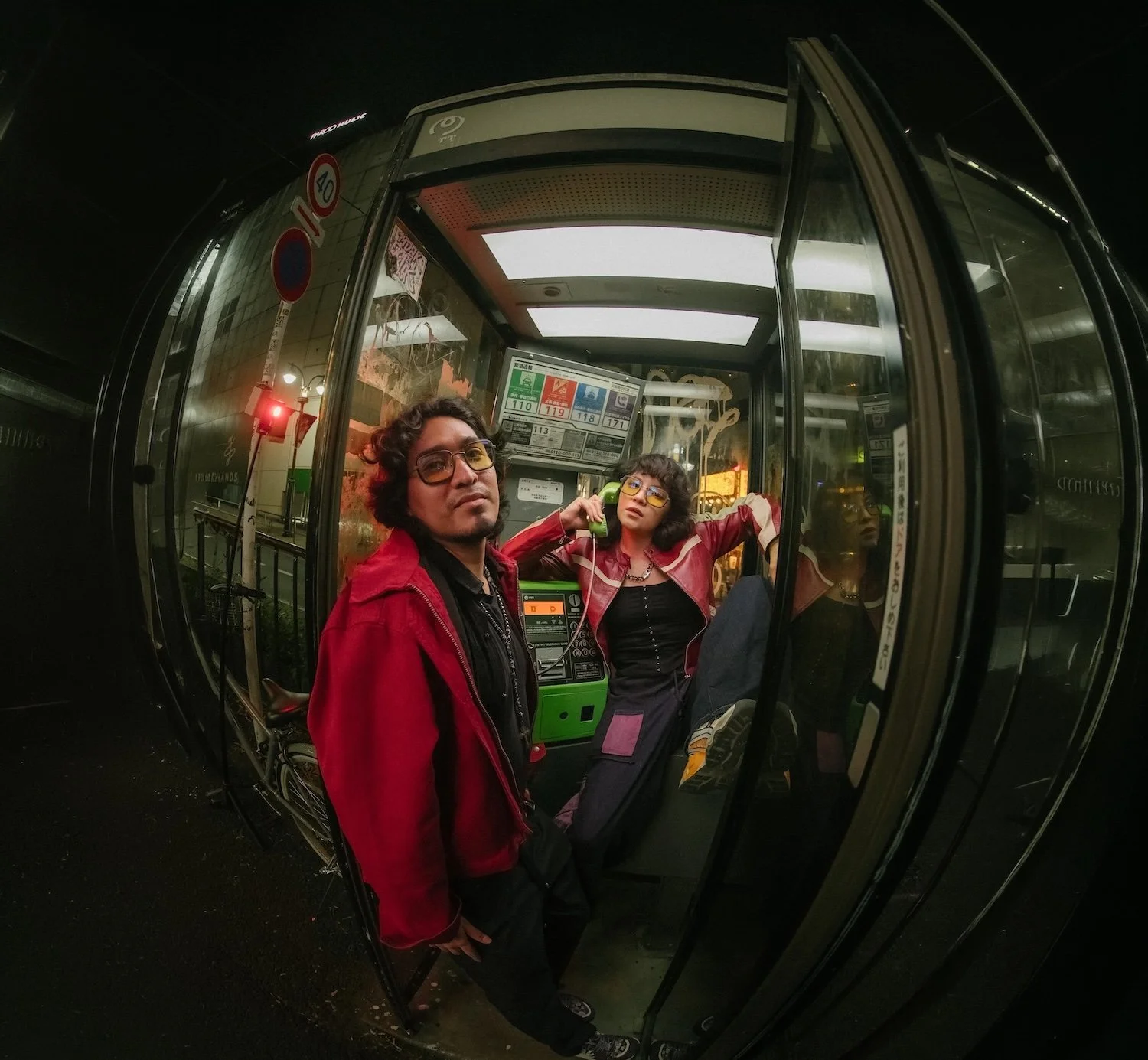 Two people inside a phone booth at night. The person on the left is a man with curly hair, glasses, and a red jacket. The person on the right is a woman with curly hair, glasses, and a pink jacket, holding a green phone to her ear. There is a city street visible through the booth's glass, with a traffic light showing red.