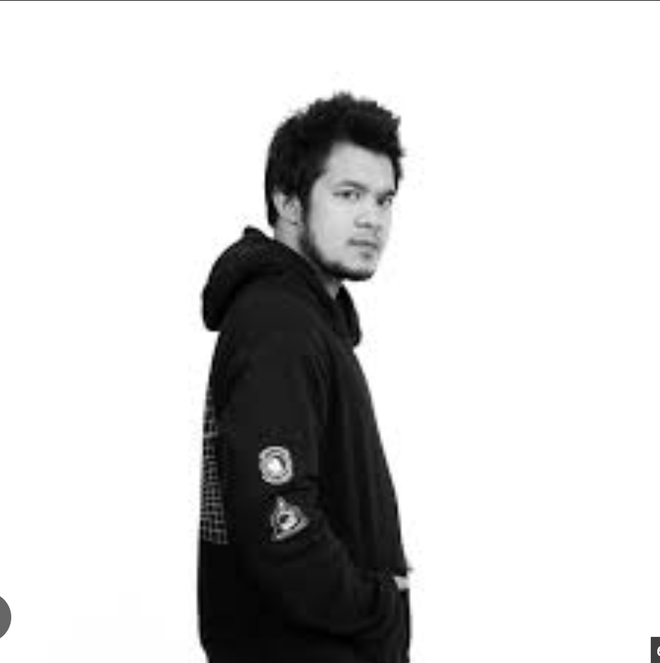 Young man with dark hair and facial hair, wearing a black hoodie with patches, standing against a plain white background.