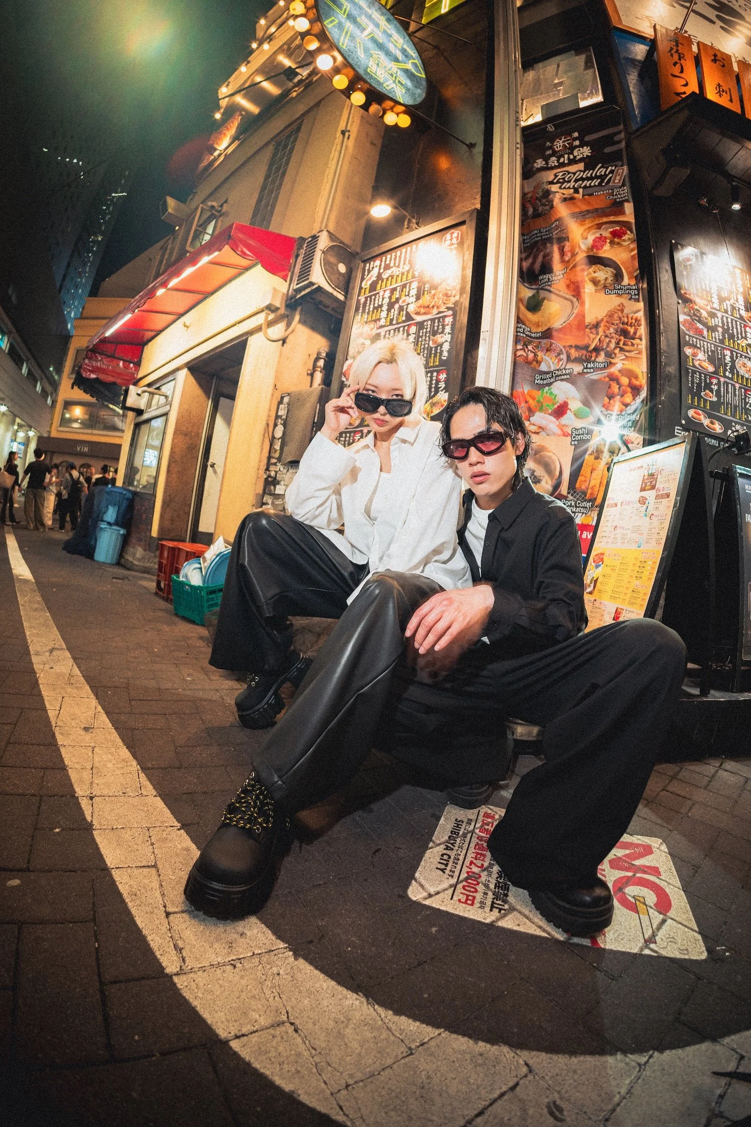 Tokyo eyewear campaign featuring Vasuma Eyewear sunglasses, with two fashion models posing outside a restaurant storefront surrounded by colorful city lights and reflections.