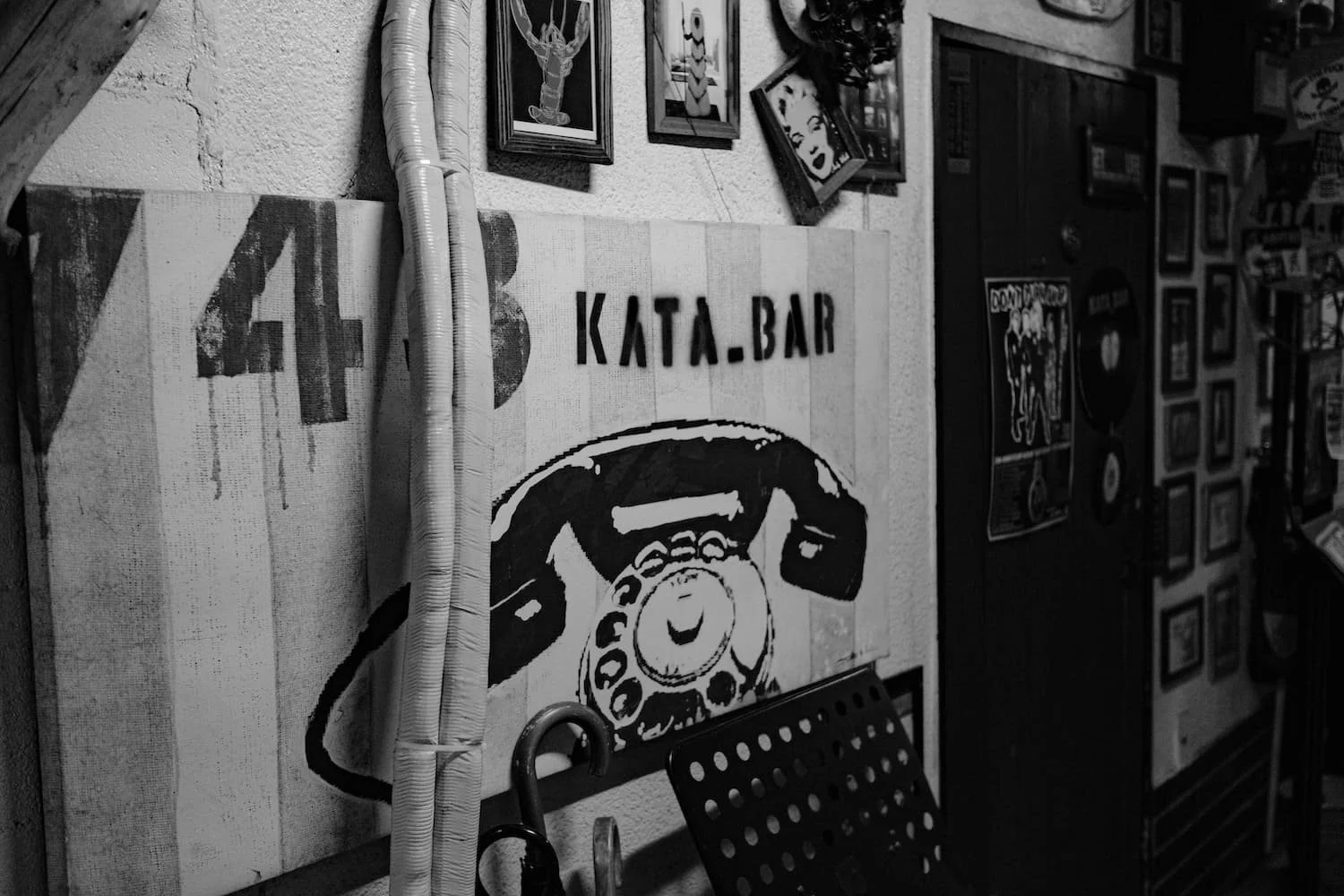 Black and white photo of a retro-style artwork of a telephone hanging on a wall at KATA.BAR, with framed pictures and posters surrounding it, and some pipes visible.