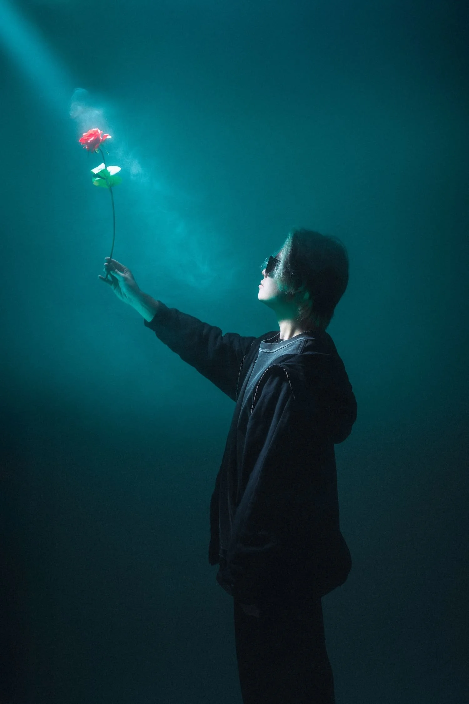 A person with dark hair and sunglasses holds a glowing, red artificial flower in a dark, teal-lit environment shot as part of a personal branding project in Tekumi Studios in Tokyo, Japan.