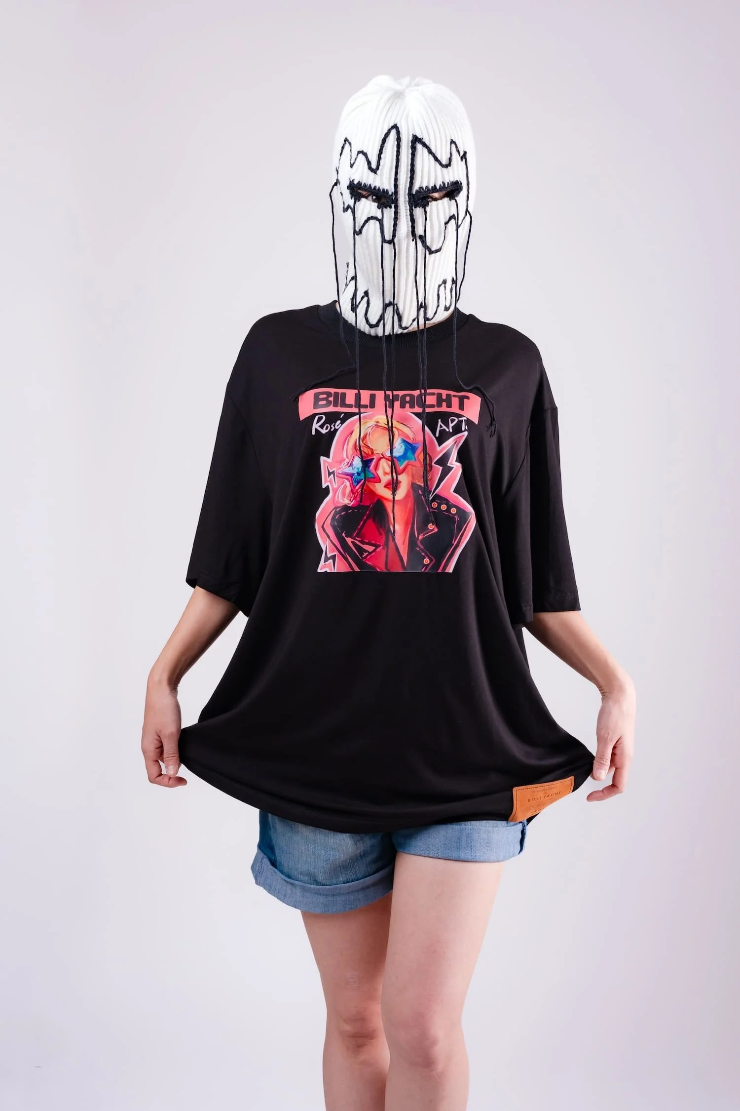 Studio portrait of a masked model wearing an oversized graphic t-shirt featuring a neon pop illustration, posing against a bright white background, photographed by Tokyo photographer Deniz Demir.
