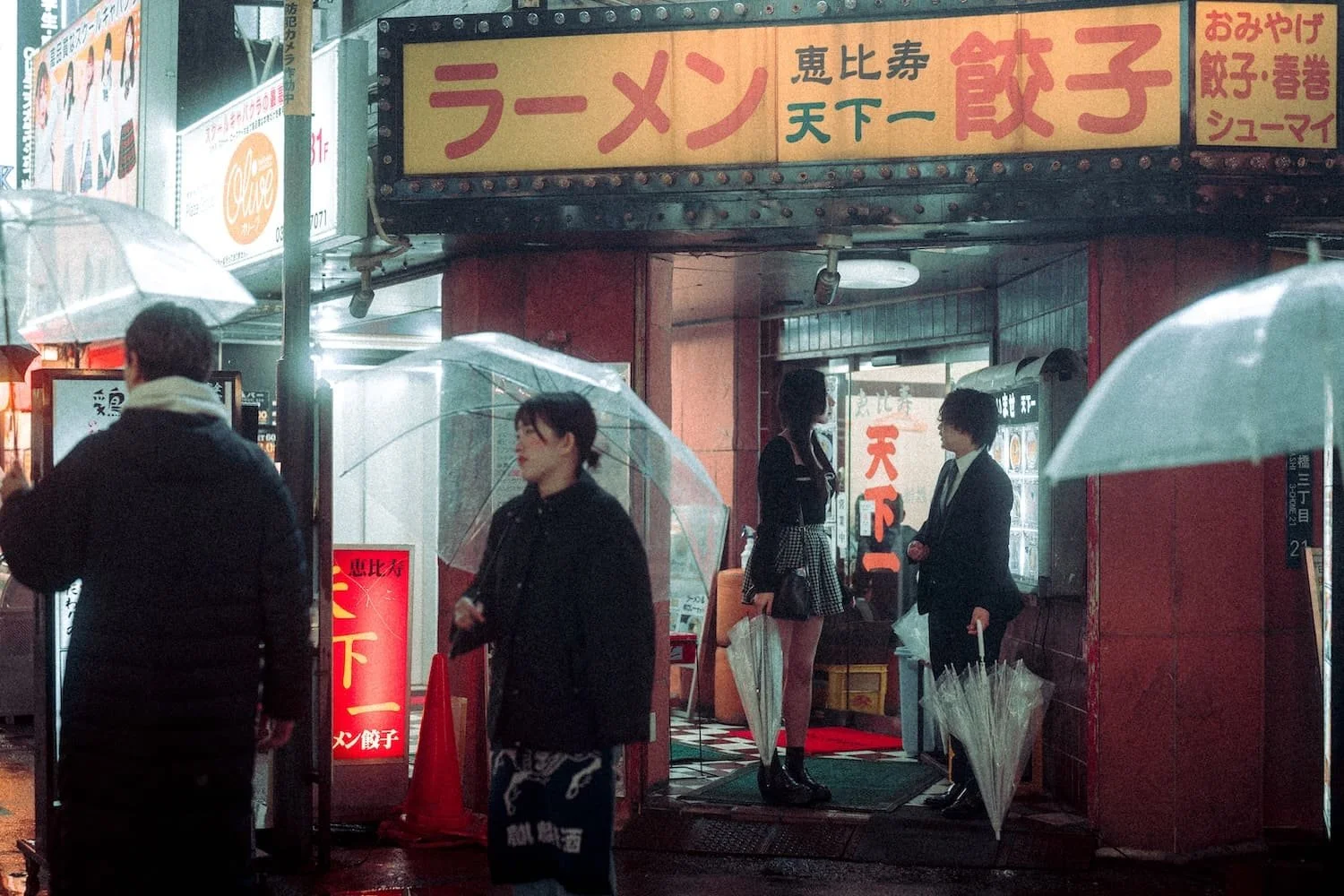 People with umbrellas standing in front of a ramen restaurant on a rainy night in Japan, with signs displaying Japanese characters