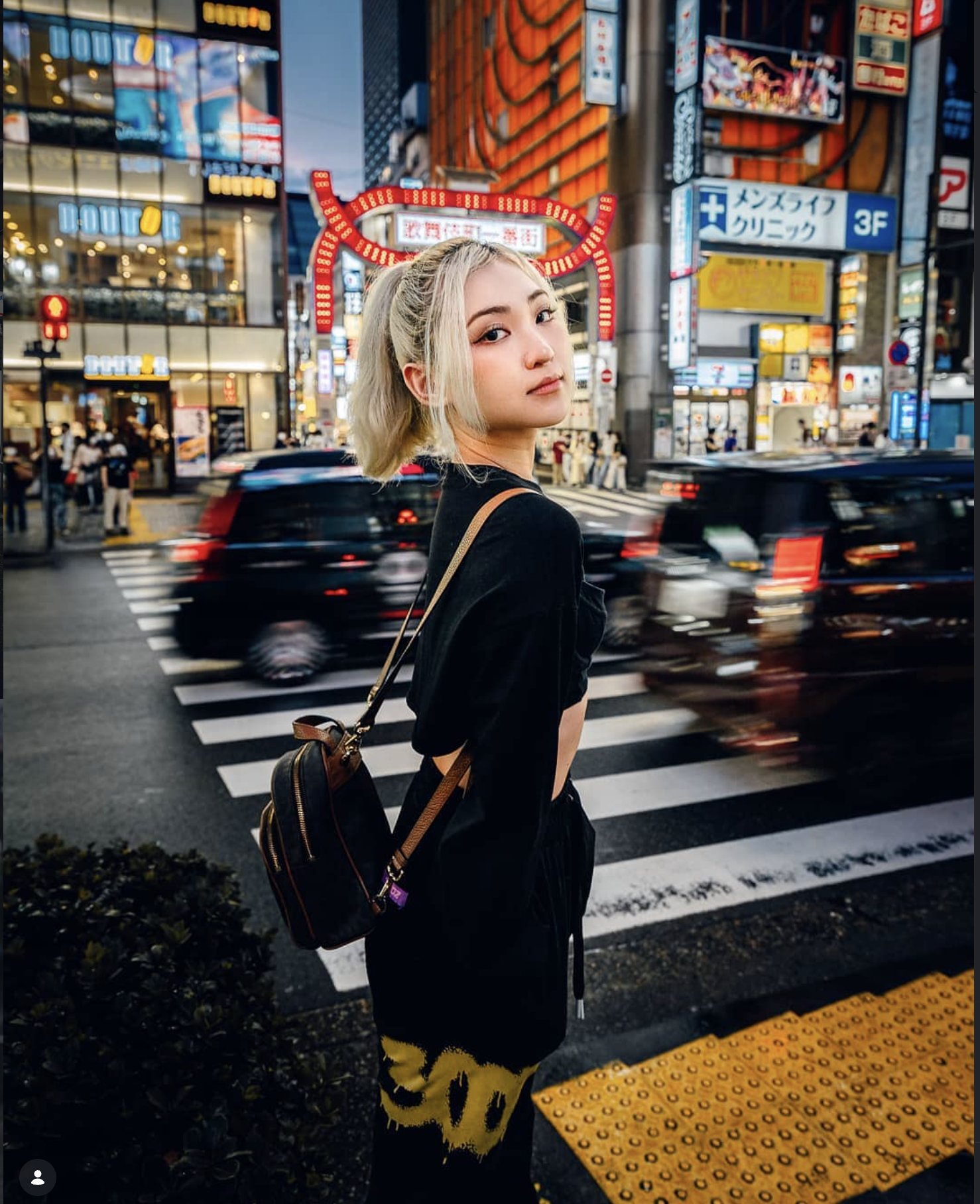A young woman with platinum blonde hair styled in a half-up, half-down look stands on a busy city street at dusk, with illuminated signs and storefronts behind her. She wears a black top and patterned yellow pants, carrying a brown backpack with black accents, and looks over her shoulder at the camera.