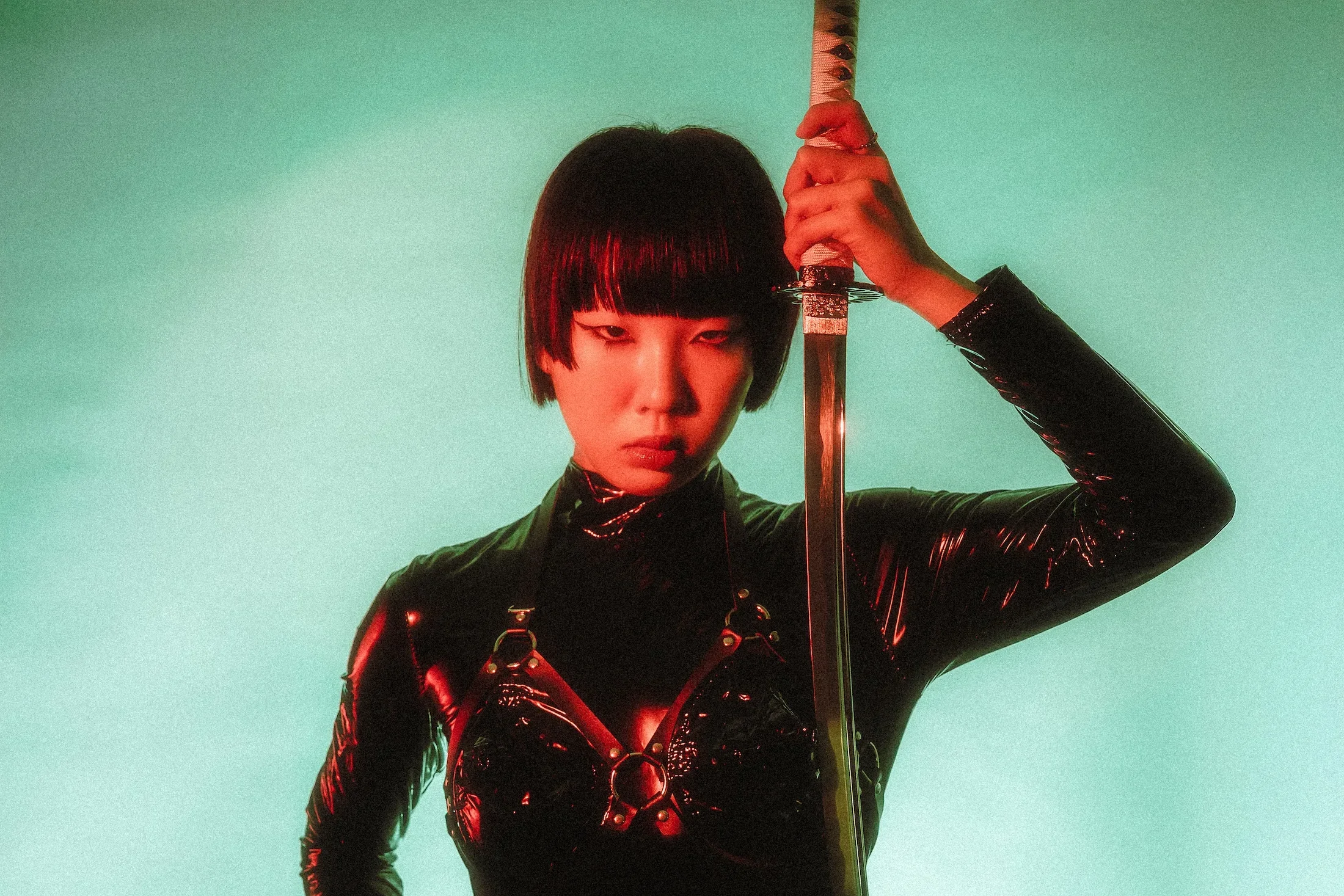 A woman with short black hair holding a sword upright in front of her face, wearing a shiny black outfit against a green background.
