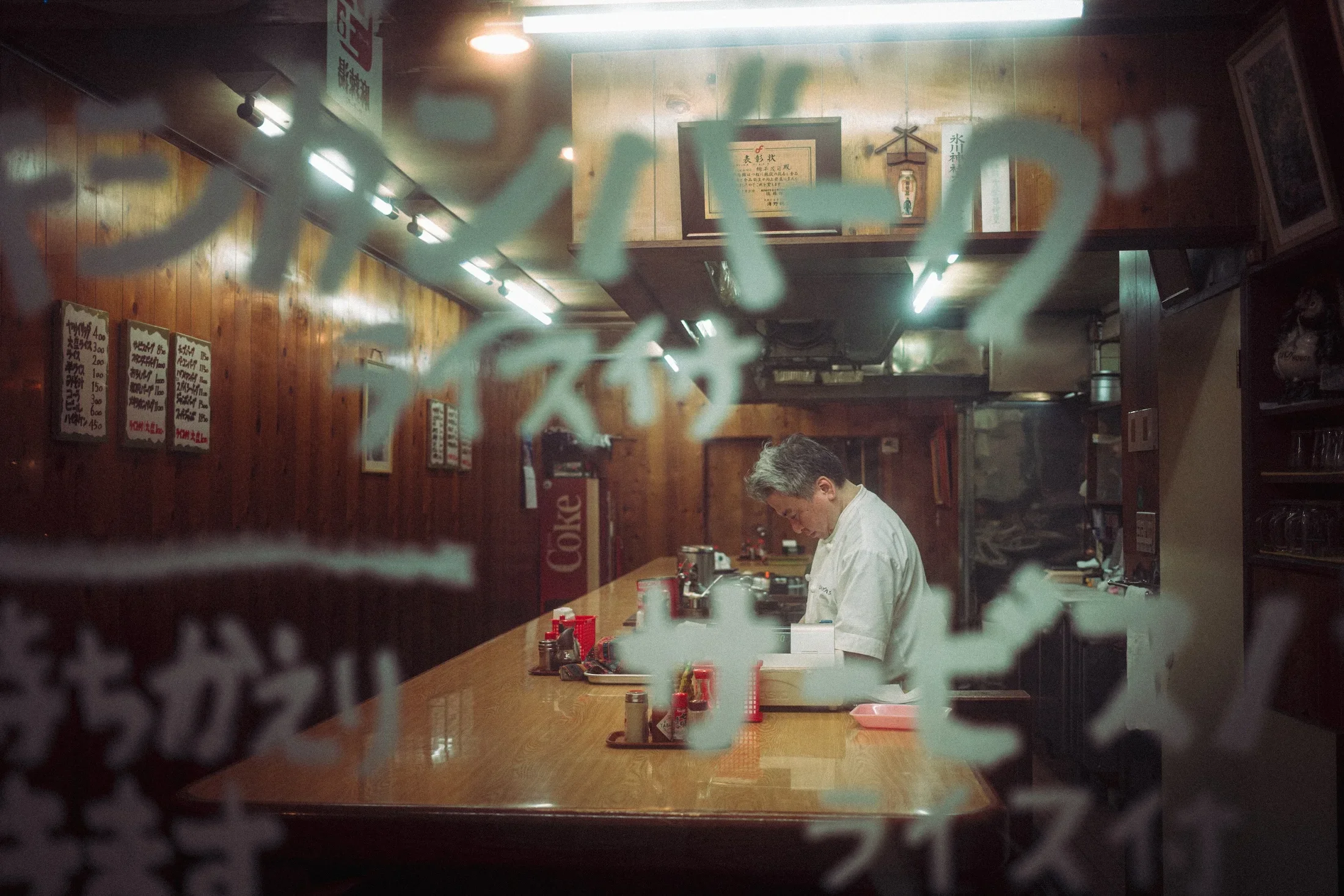 An interior view of a Japanese restaurant seen through a decorative window pane with Japanese characters in the foreground. The restaurant has a wooden wall with menu signs on the left, framed papers and a small bottle decoration on the wall in the background. A man in a white shirt is working behind the counter.