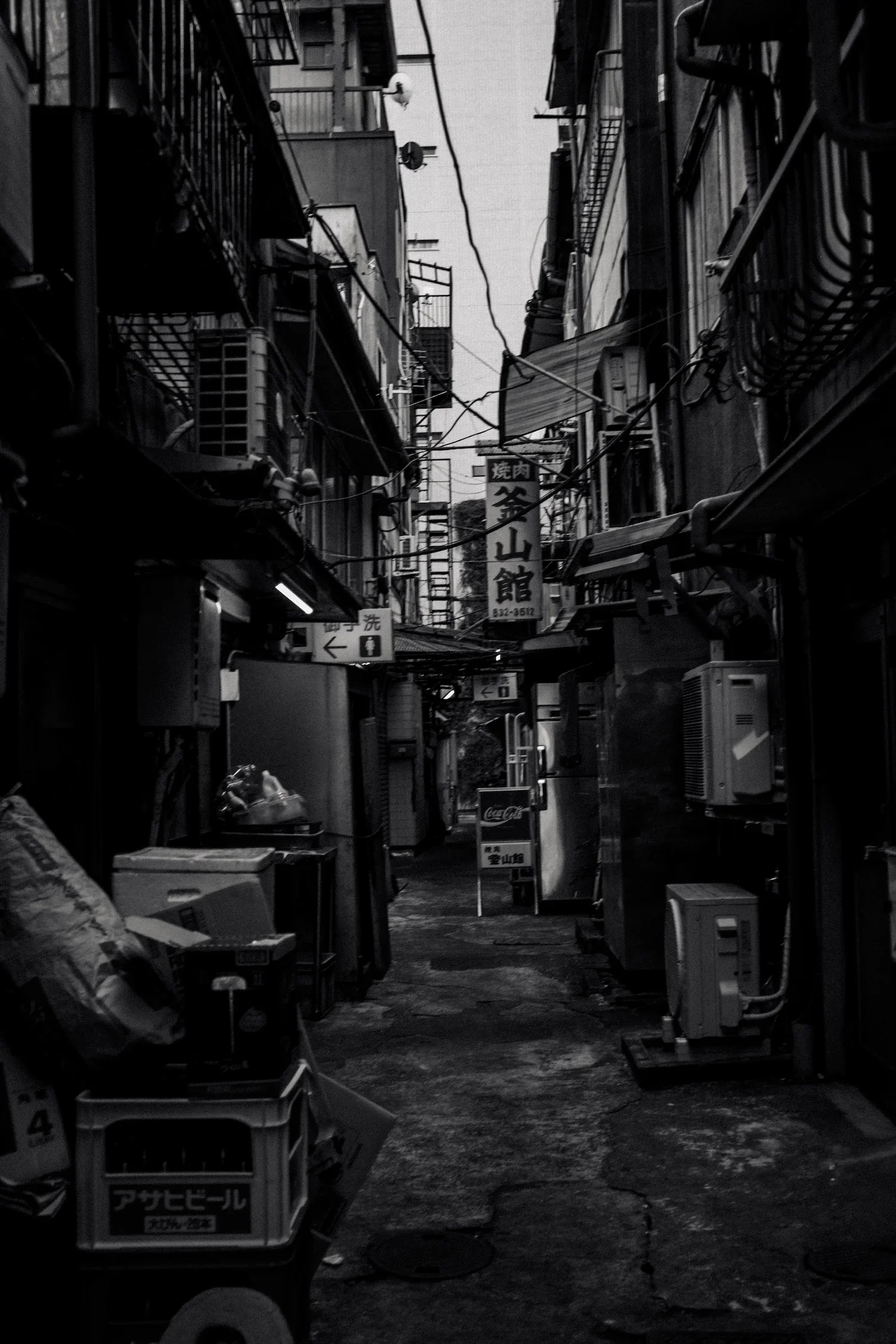 A narrow alleyway in an urban area with various signs, electrical wires, air conditioning units, and clutter on the ground. The photo is in black and white.