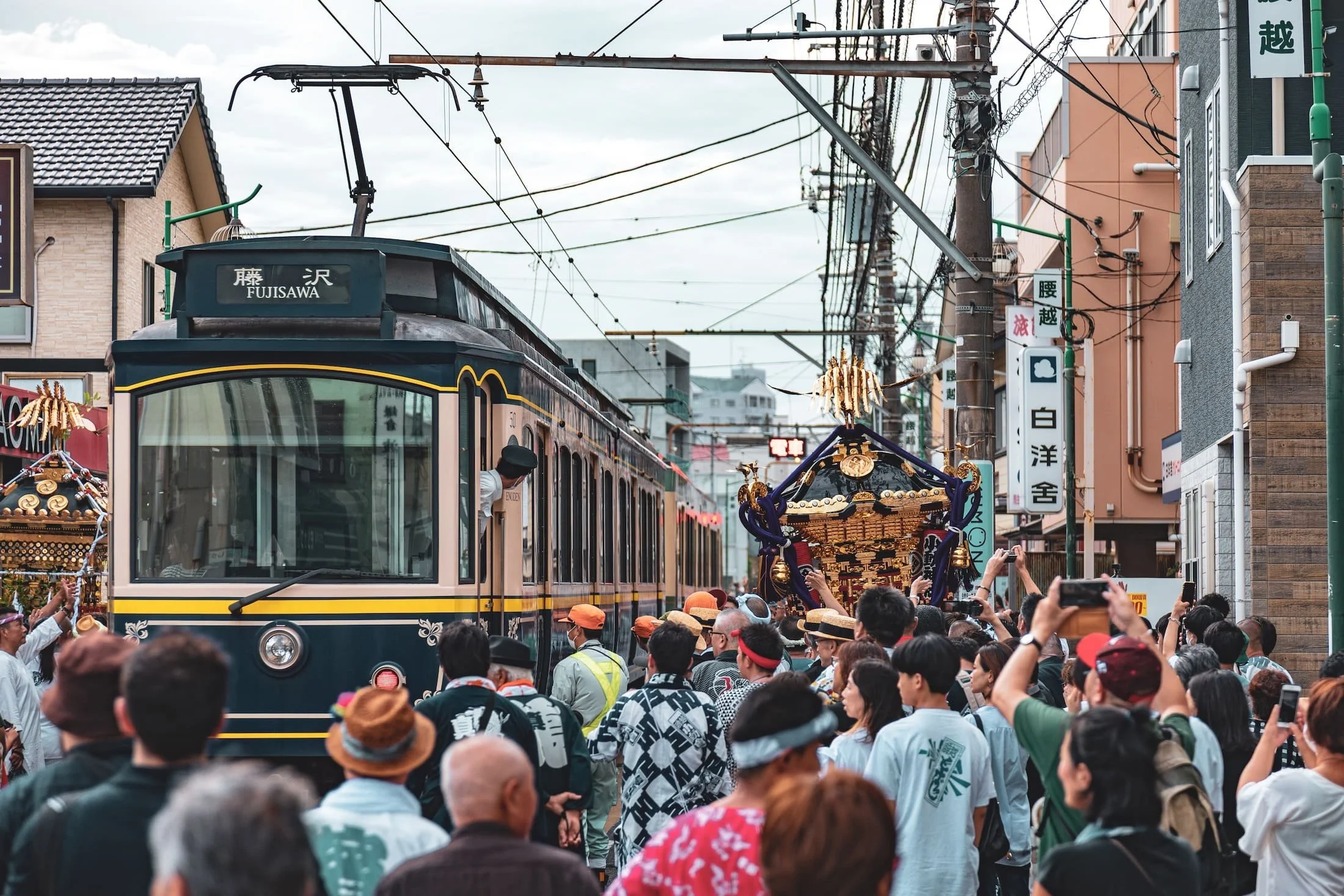 A busy street scene during a festival with a crowd of people, a tram, traditional festival elements, and electrical wires overhead.
