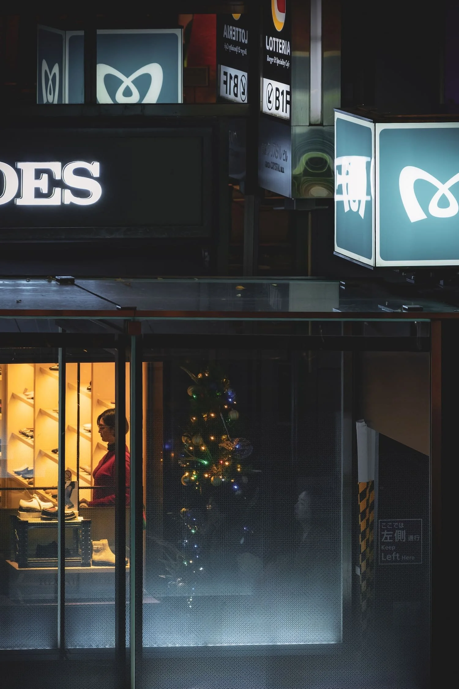 A woman shopping inside a store decorated with a small Christmas tree with lights, viewed through a glass window at night.