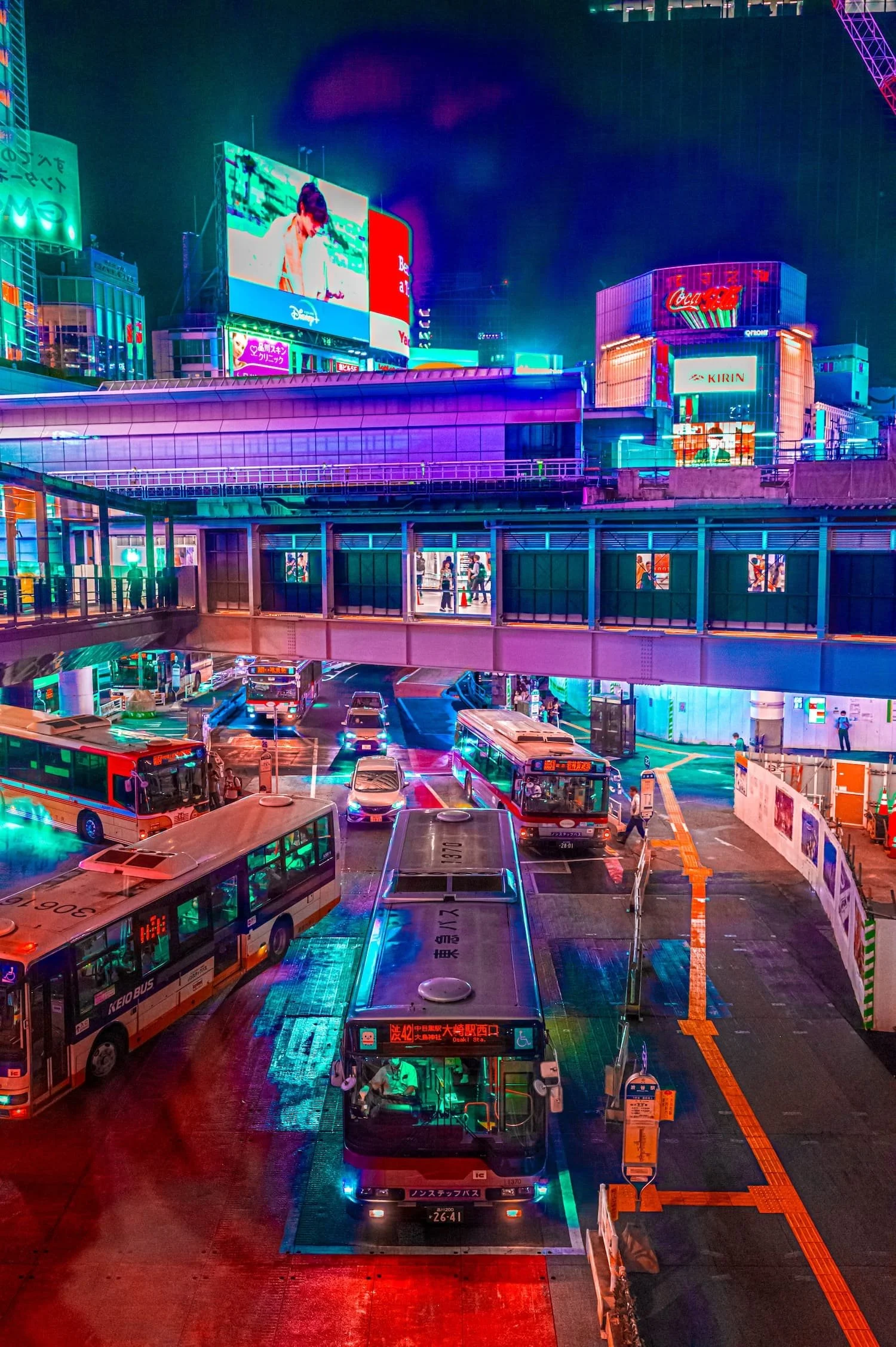 City street at night illuminated with neon lights, featuring buses and cars at a bus stop with a pedestrian bridge overhead, surrounded by vibrant digital billboards and advertisements.
