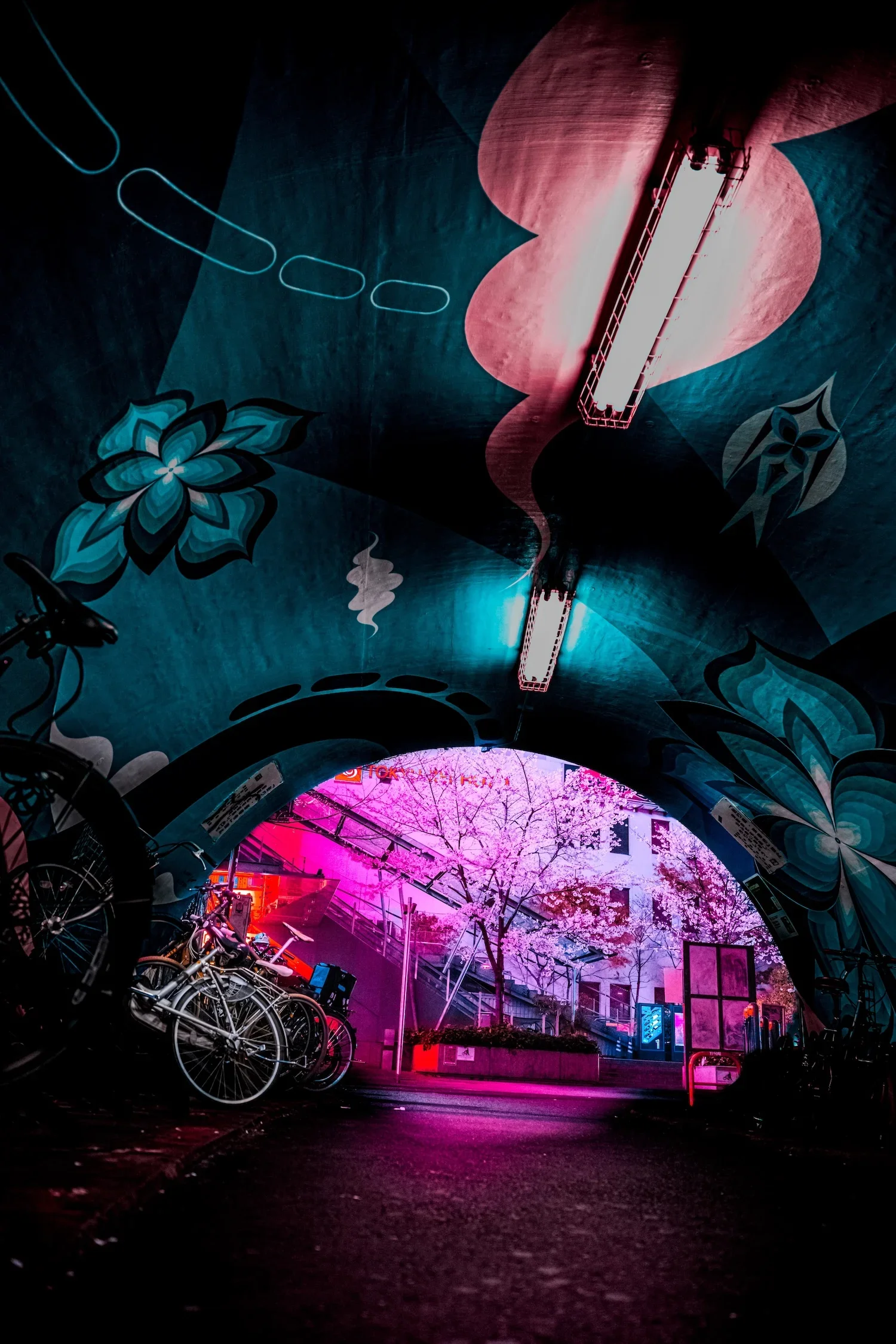 Nighttime city scene viewed from underneath an arched tunnel with colorful murals and bicycles parked nearby, pink lighting reflecting on the ground.