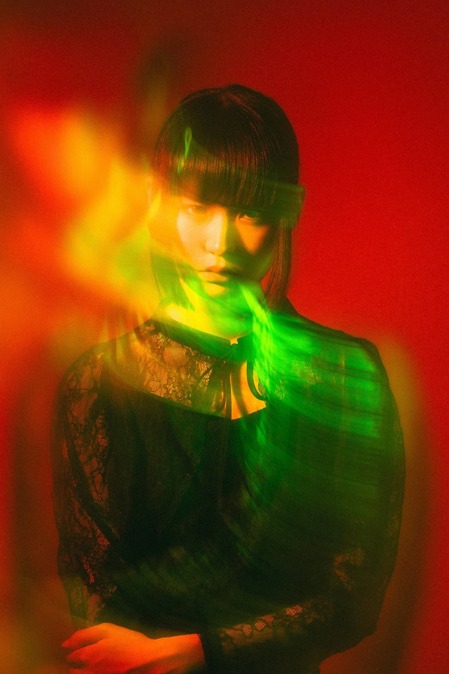 A woman with straight black hair and bangs wearing a black lace top, illuminated by yellow and green light effects in a red background.