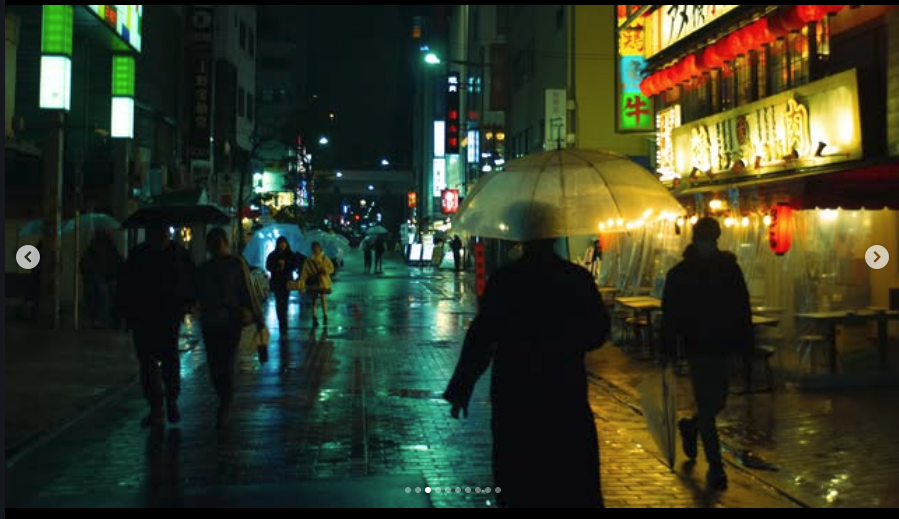 City street at night with wet pavement, illuminated signs, and people walking with umbrellas.