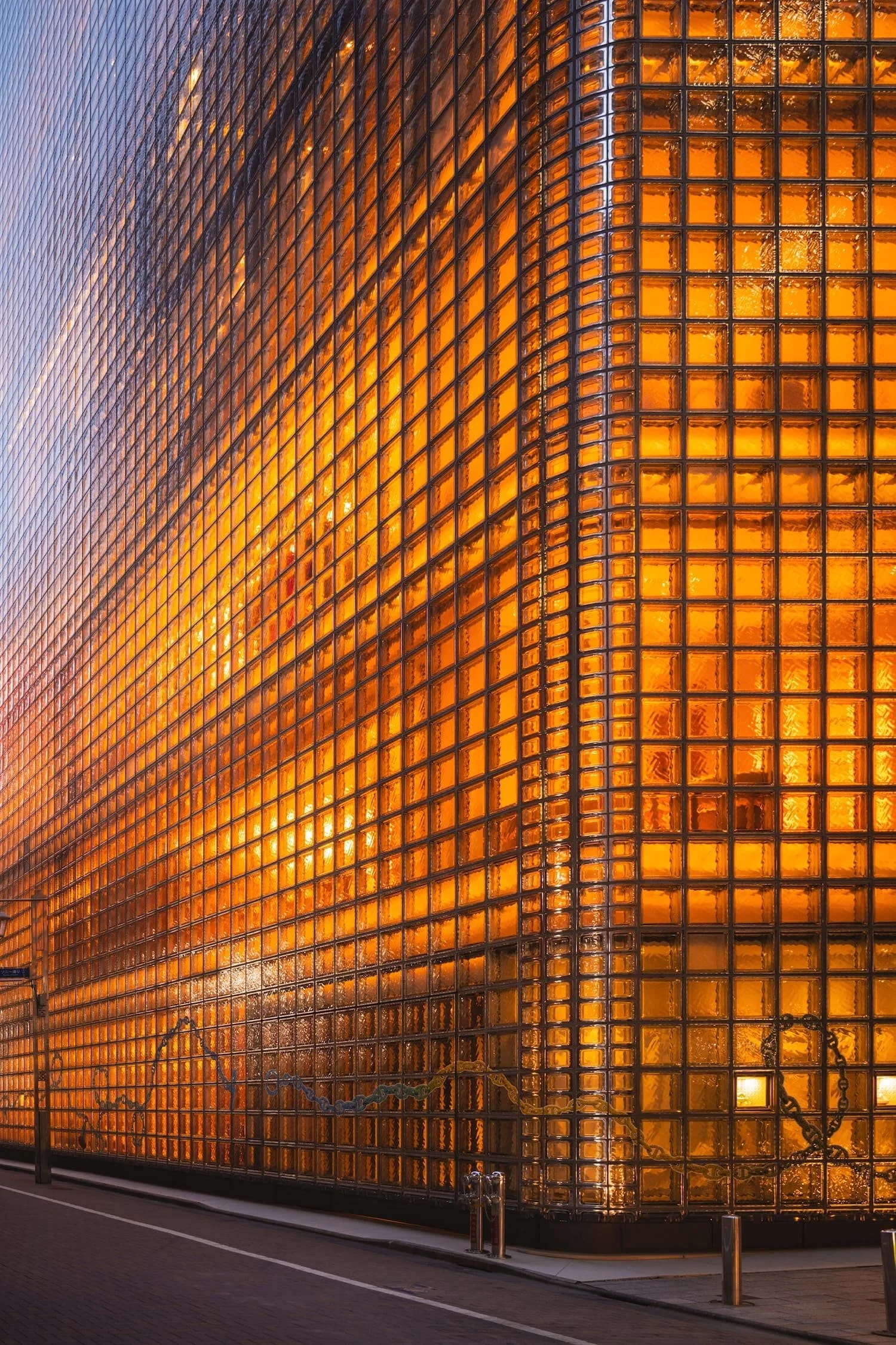 A modern glass building with a unique orange-tinted glass façade reflecting the sky, illuminated from inside, with a sidewalk and street in front.