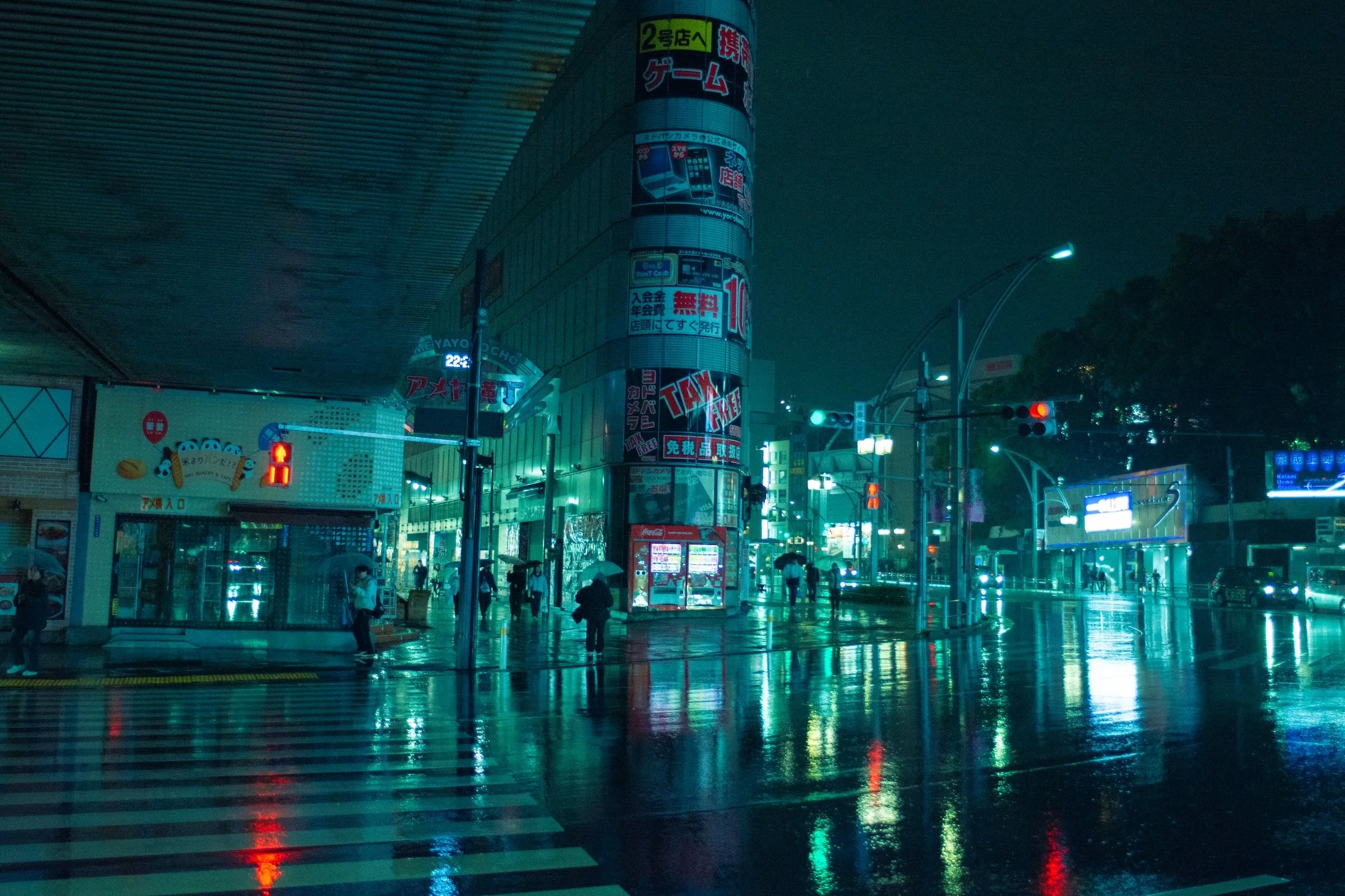 Nighttime city street scene with illuminated signs in Japanese, wet pavement reflecting lights, and a few people holding umbrellas.