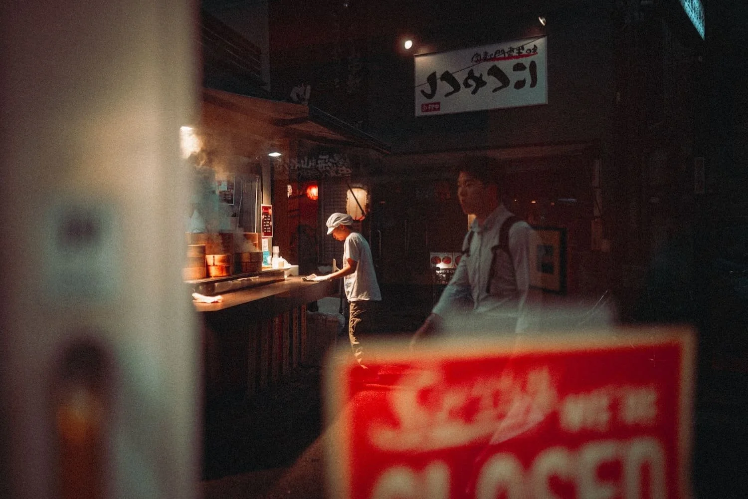 Inside a dimly lit restaurant with two staff members visible, one is a woman in a white cap working at a counter, and the other is a man in a white shirt with a backpack, standing nearby. There is a sign with foreign characters hanging from the ceili