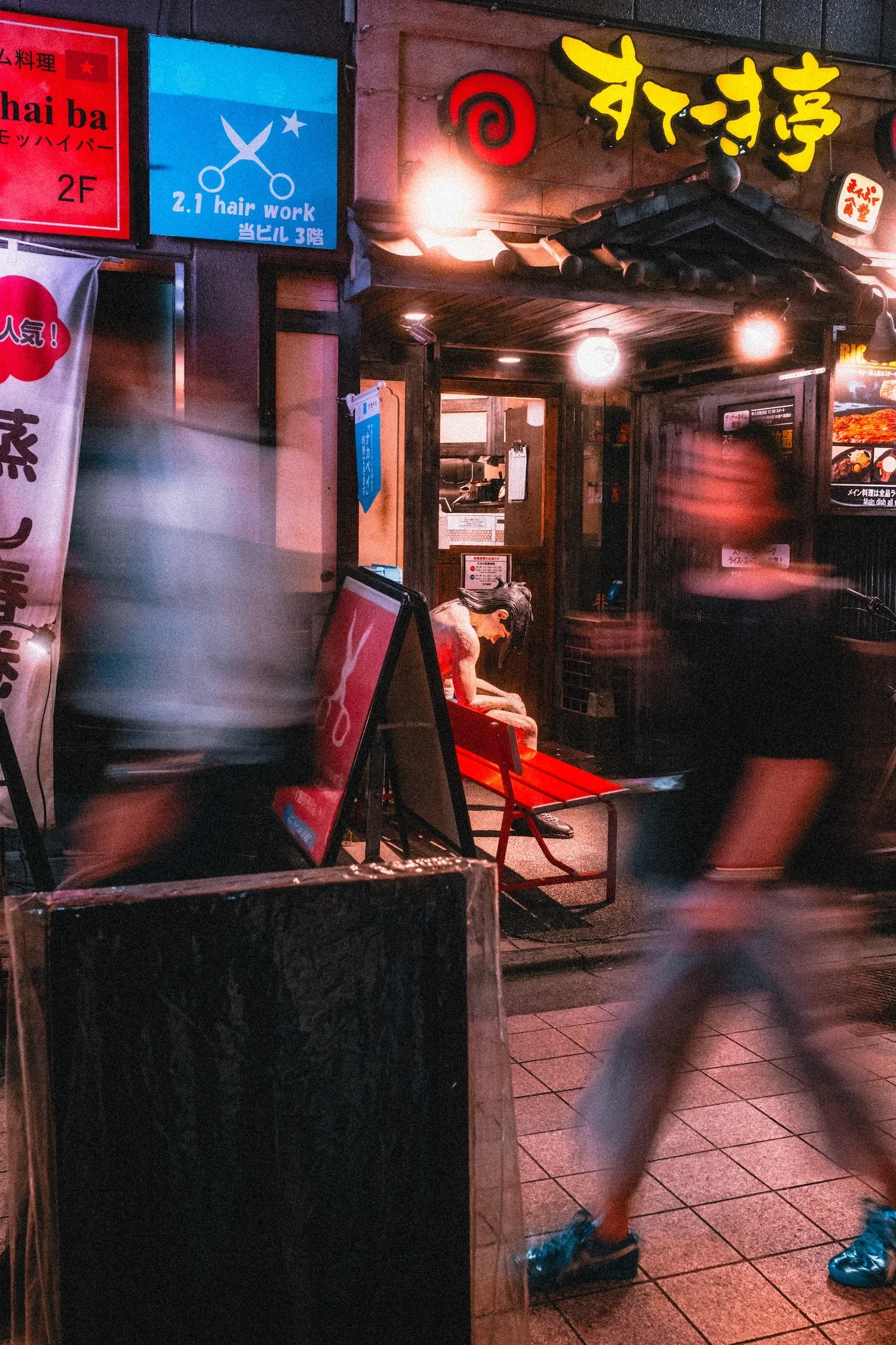 Nighttime street scene showing the entrance of a Japanese restaurant with illuminated signs in yellow and red. A person is sitting on a small red bench outside, with motion blur indicating movement. There are various signs in Japanese and English advertising local businesses, including a hair salon and possibly other shops. The scene is lively with blurred motion of pedestrians passing by.