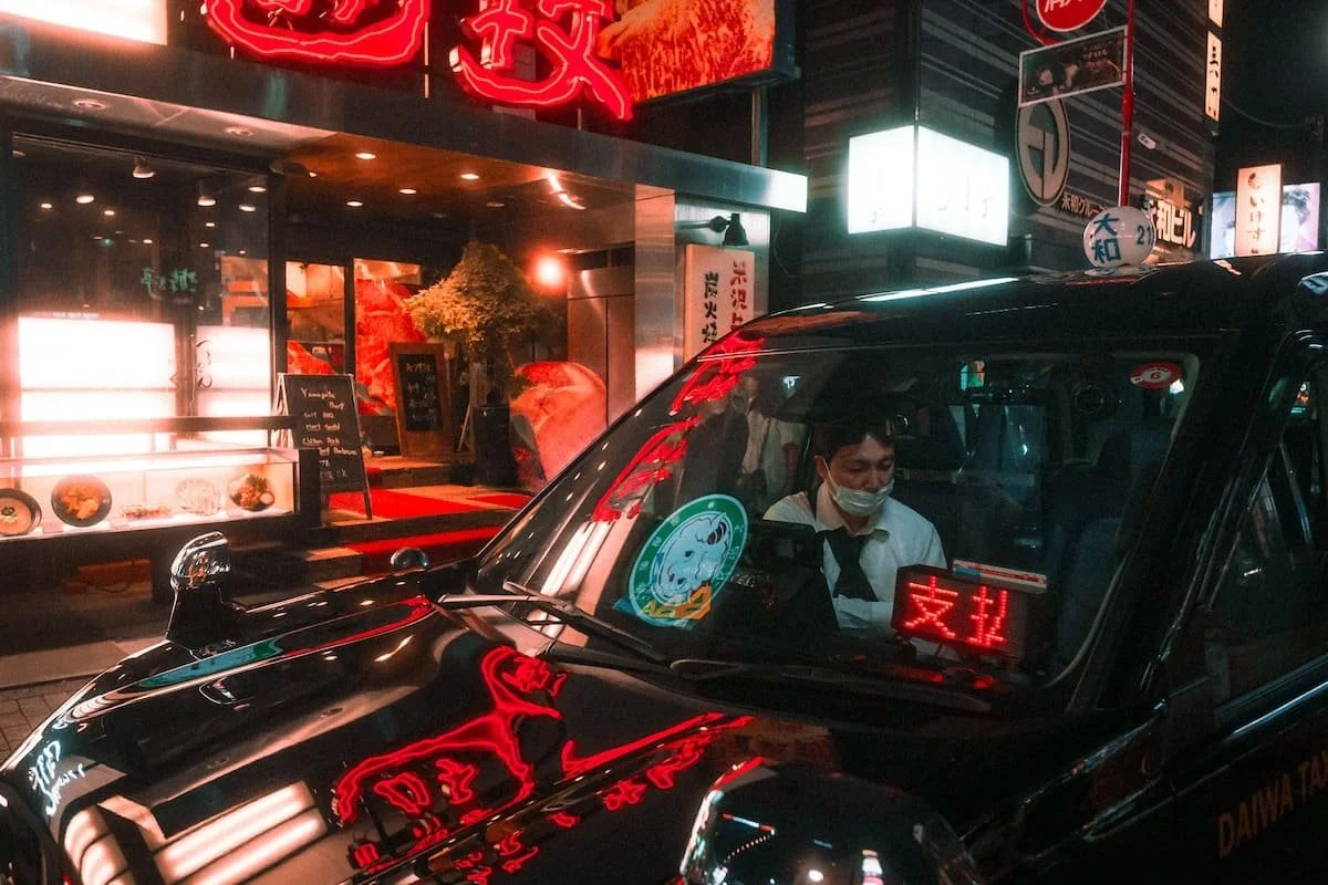 A taxi driver wearing a face mask sitting inside a black taxi with illuminated signs in a neon-lit city background at night.