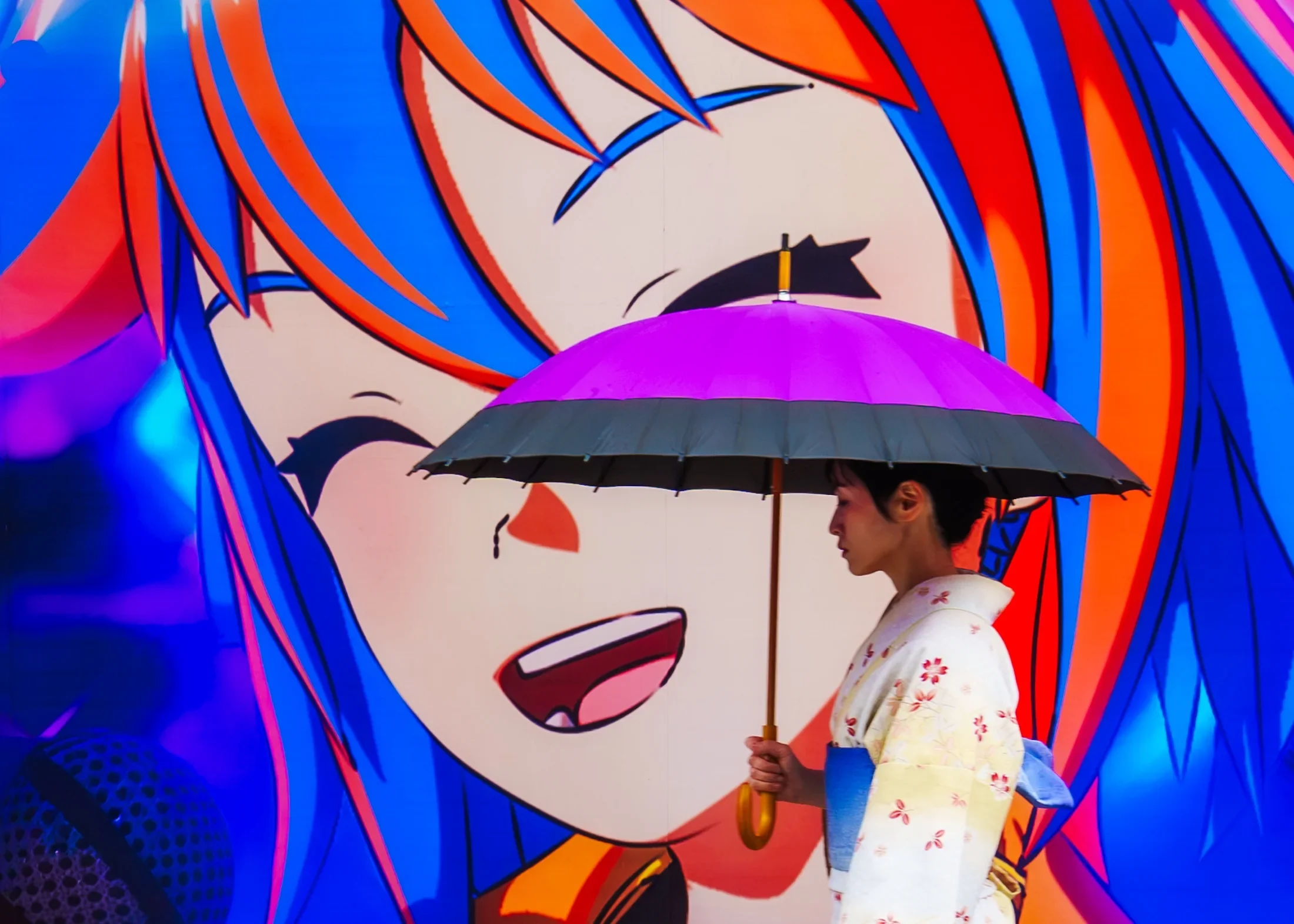 A person holding a purple and black umbrella stands in front of a colorful graffiti mural of a woman with multicolored hair, pink lips, and black eye makeup.