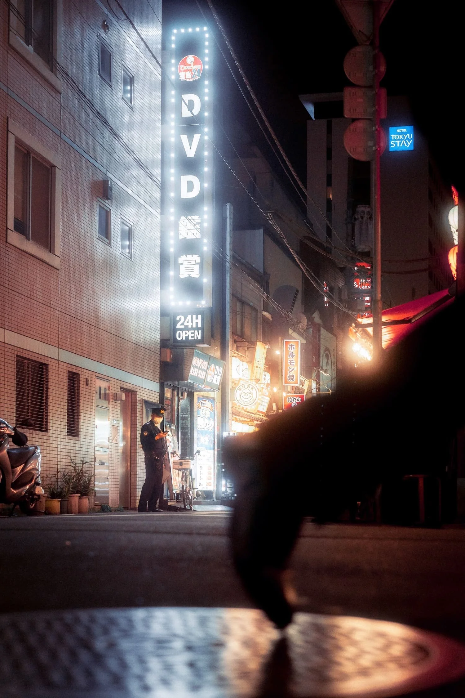 A nighttime street scene with illuminated signs, including a vertical sign reading 'DVD' and '24H OPEN,' a police officer on the sidewalk looking at a device, and various other neon signs in a city area shot by Tokyo photographer Deniz Demir.