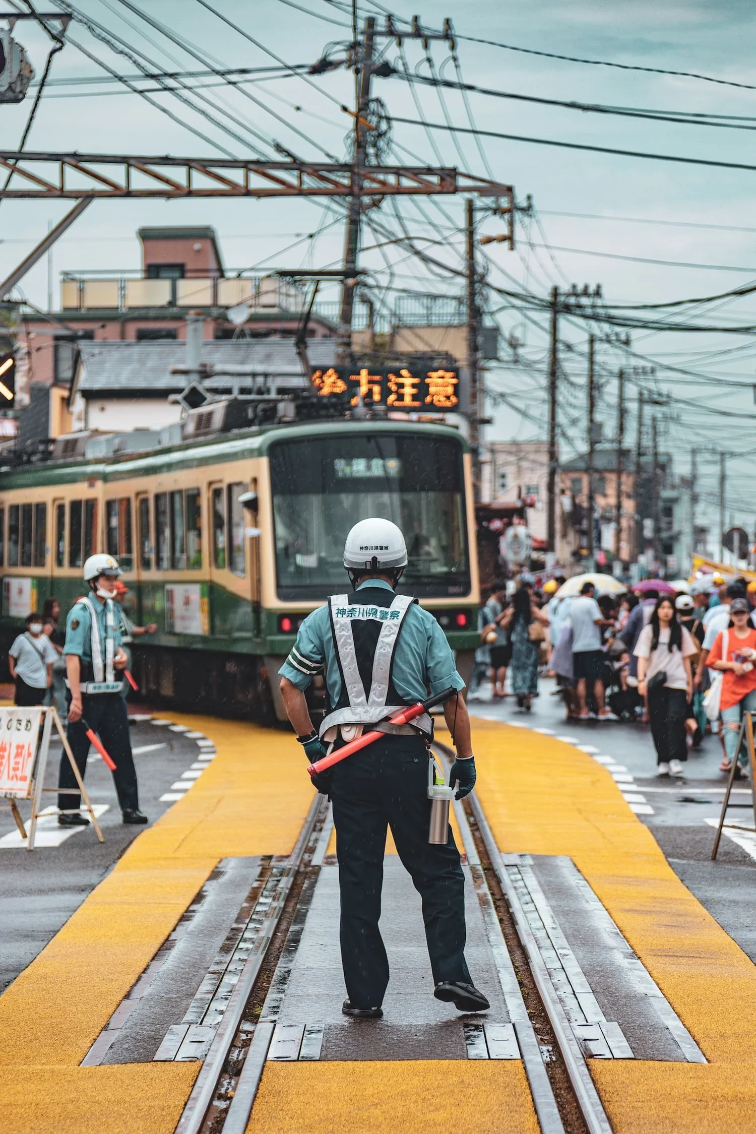 A police officer standing on a train crossing marked with yellow tactile paving, directing a passing train on a busy street full of pedestrians and power lines in a Japanese urban setting.