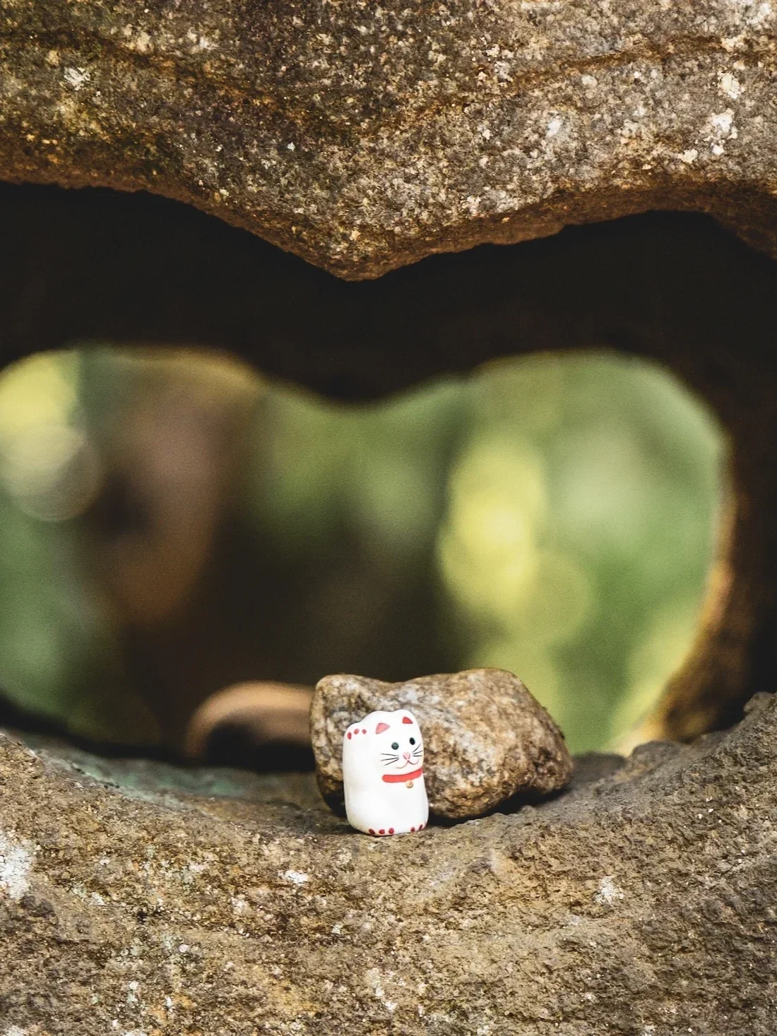Small ceramic figurine shaped like a cat with red accents, sitting on a rock with a larger rock behind it, outdoors with blurred green and brown background.