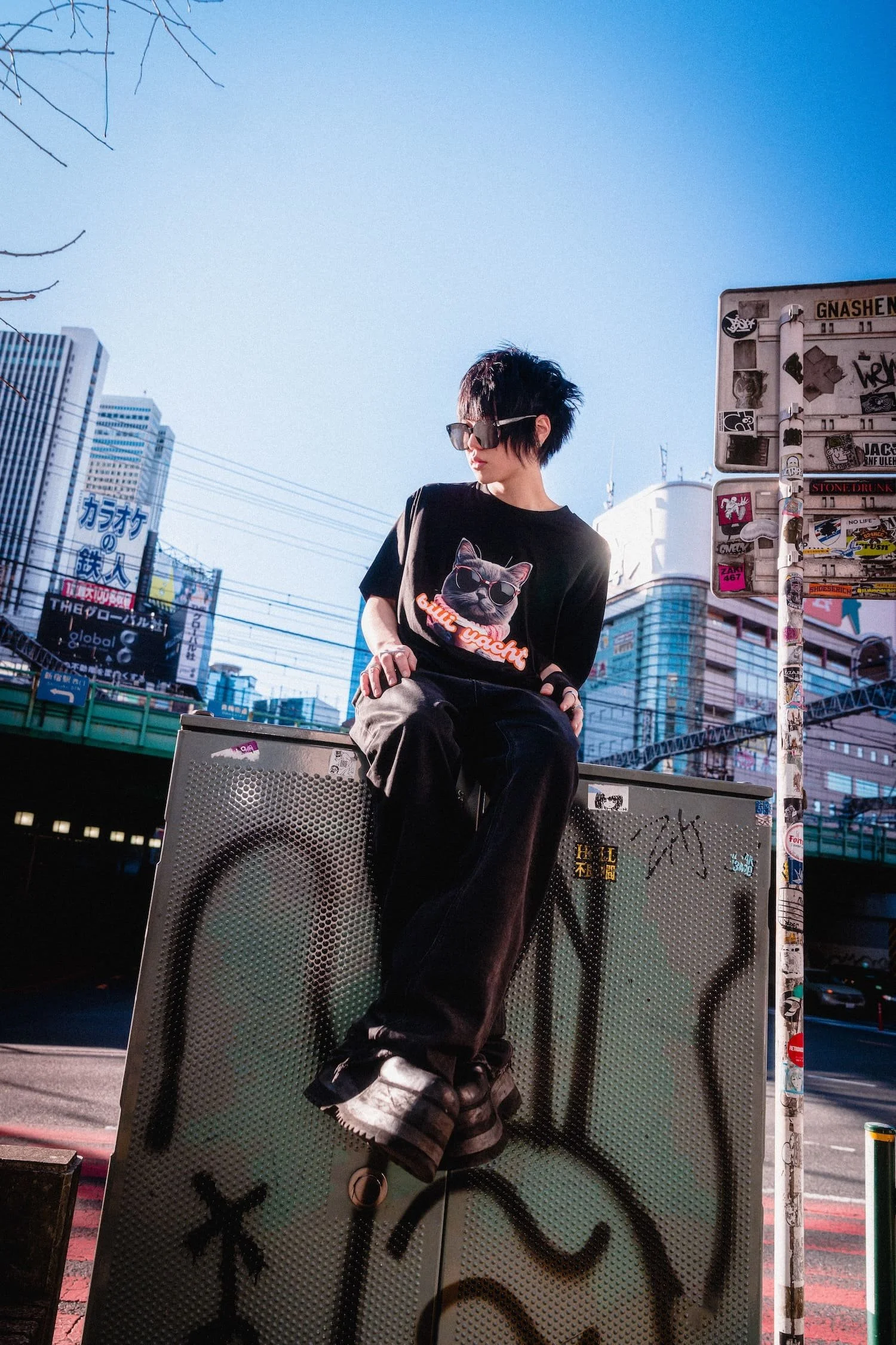 Urban fashion portrait of a model sitting atop a graffiti-covered street utility box in Tokyo, wearing sunglasses and a graphic cat t-shirt, with elevated train tracks and city buildings behind them, photographed by Tokyo photographer Deniz Demir.