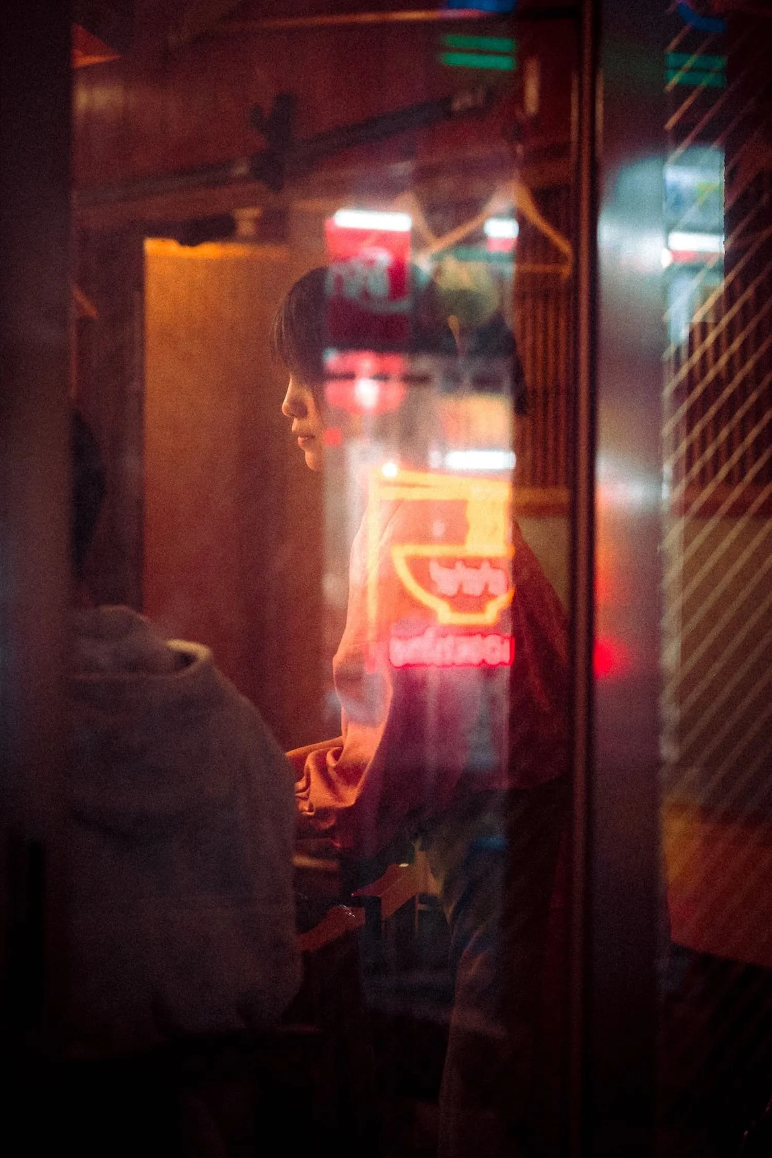 A person with dark hair is seen through a glass window with neon signs reflected on it, creating a layered, abstract scene in a dimly lit setting.