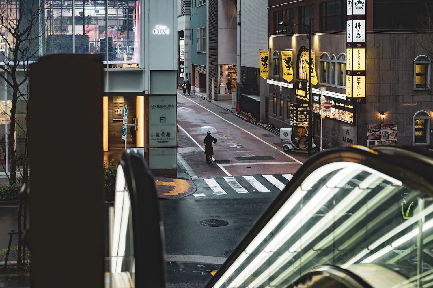 A rainy city street viewed from an elevated position, showing a person with an umbrella crossing at a crosswalk, surrounded by buildings with illuminated signs and advertisements.