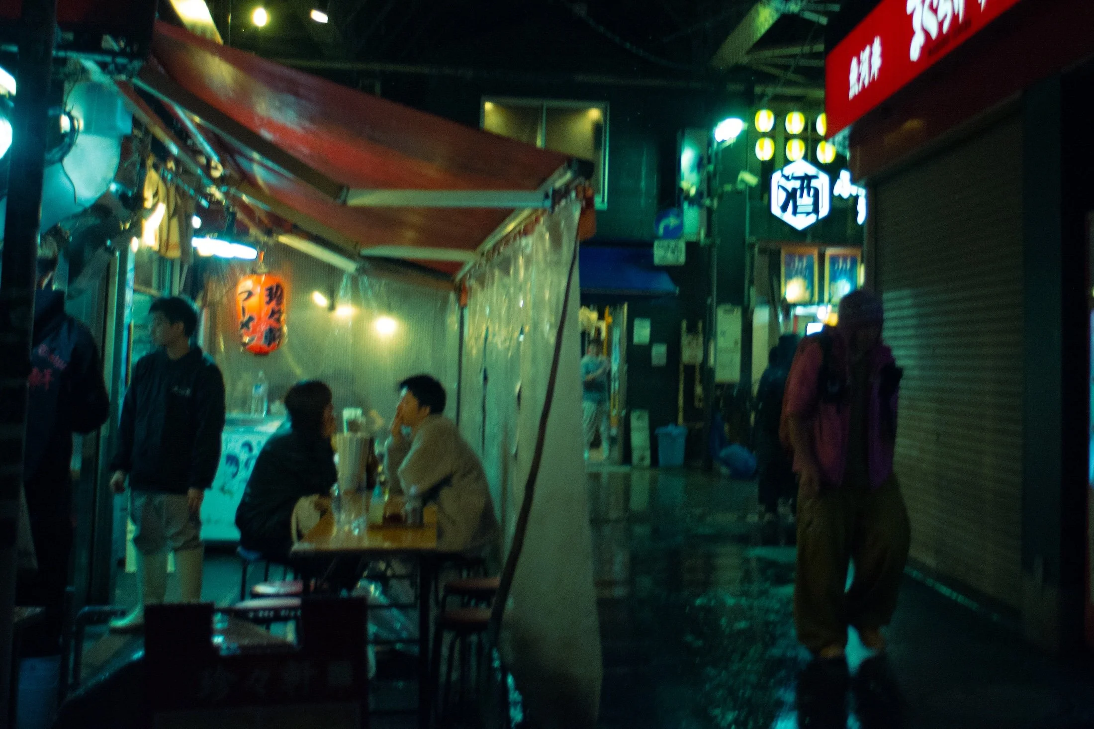 Night scene of a small outdoor market or food stall area with several people sitting at tables and others standing. Neon signs and colorful lights illuminate the scene, with some signs displaying Chinese or Japanese characters. The setting appears lively and busy.