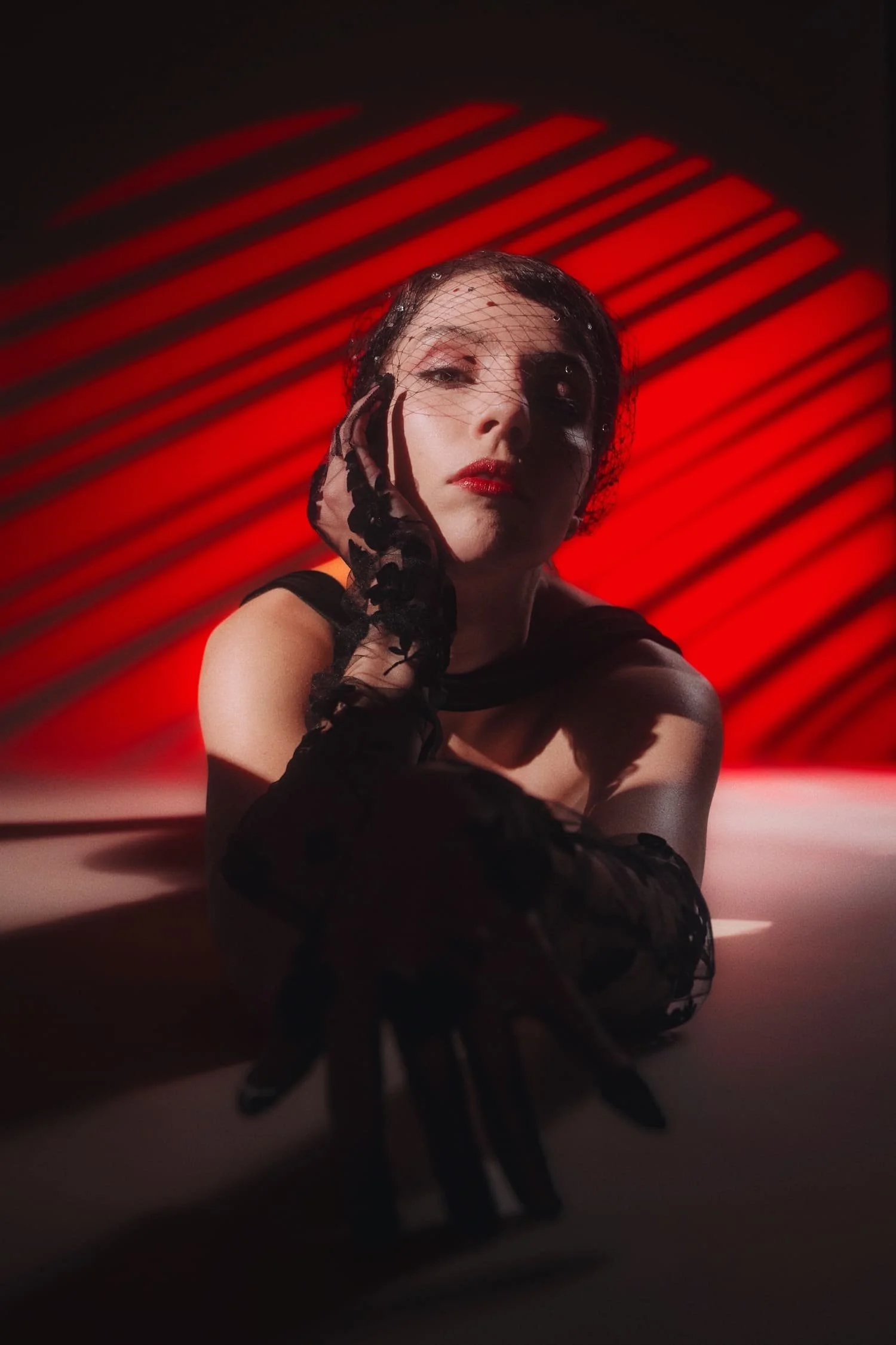 A woman with makeup and red lipstick wearing black lace gloves and a black headscarf, posing against a red and black striped background.