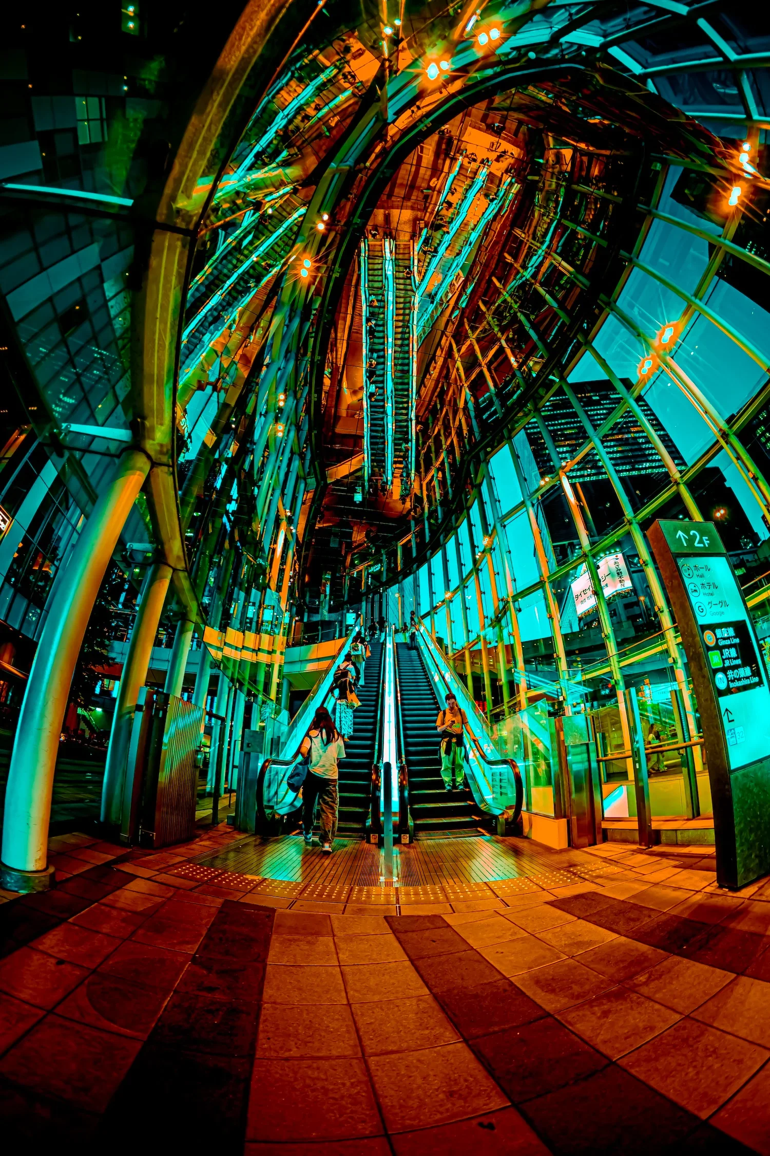 Night view of a modern, colorful architecture featuring glass and steel. A staircase with people ascending and descending is centered, illuminated by vibrant lights reflecting a futuristic atmosphere.