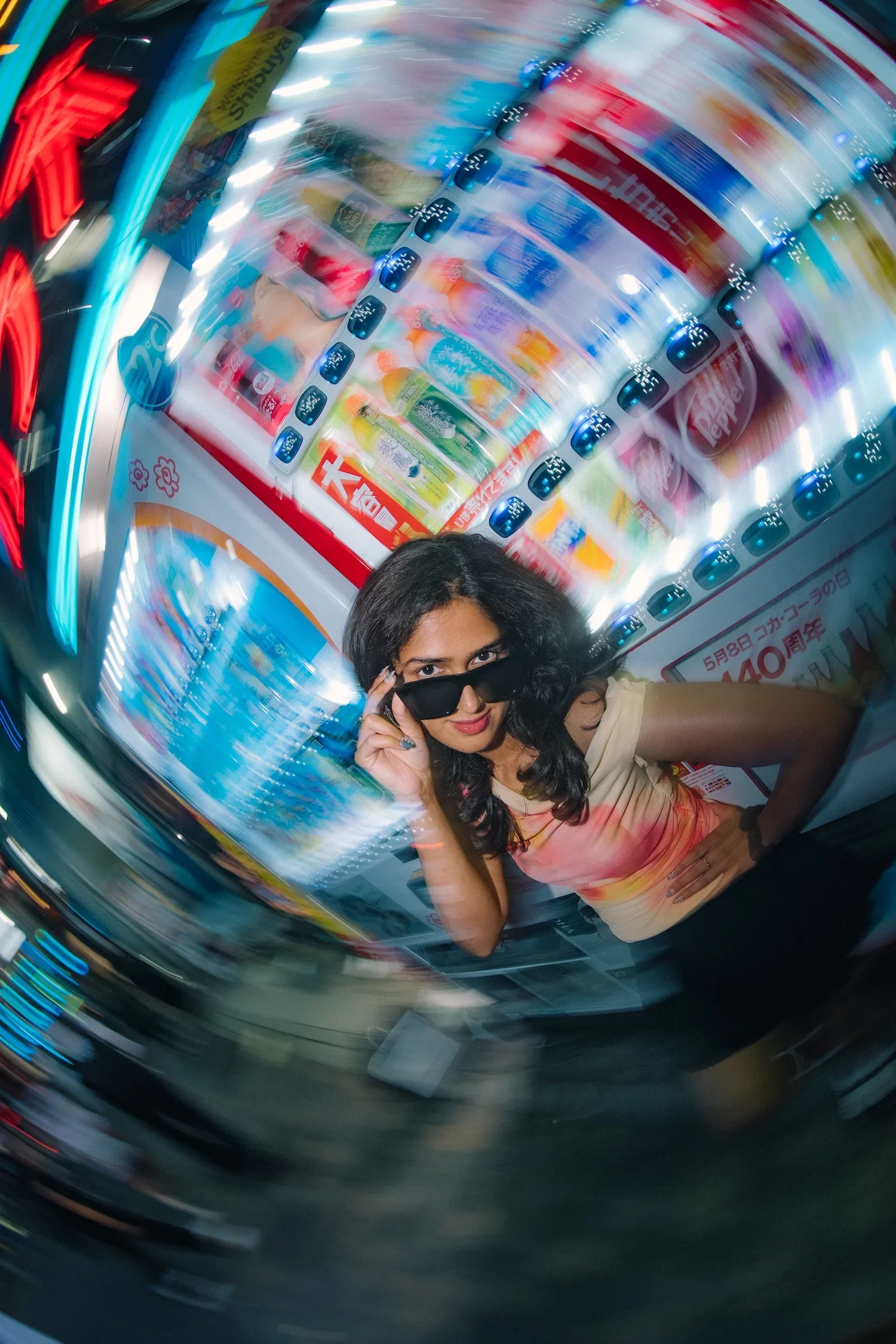 A woman with dark hair, wearing sunglasses and a beige top, standing in front of a blurry, swirling backdrop of vending machines and colorful advertisements.