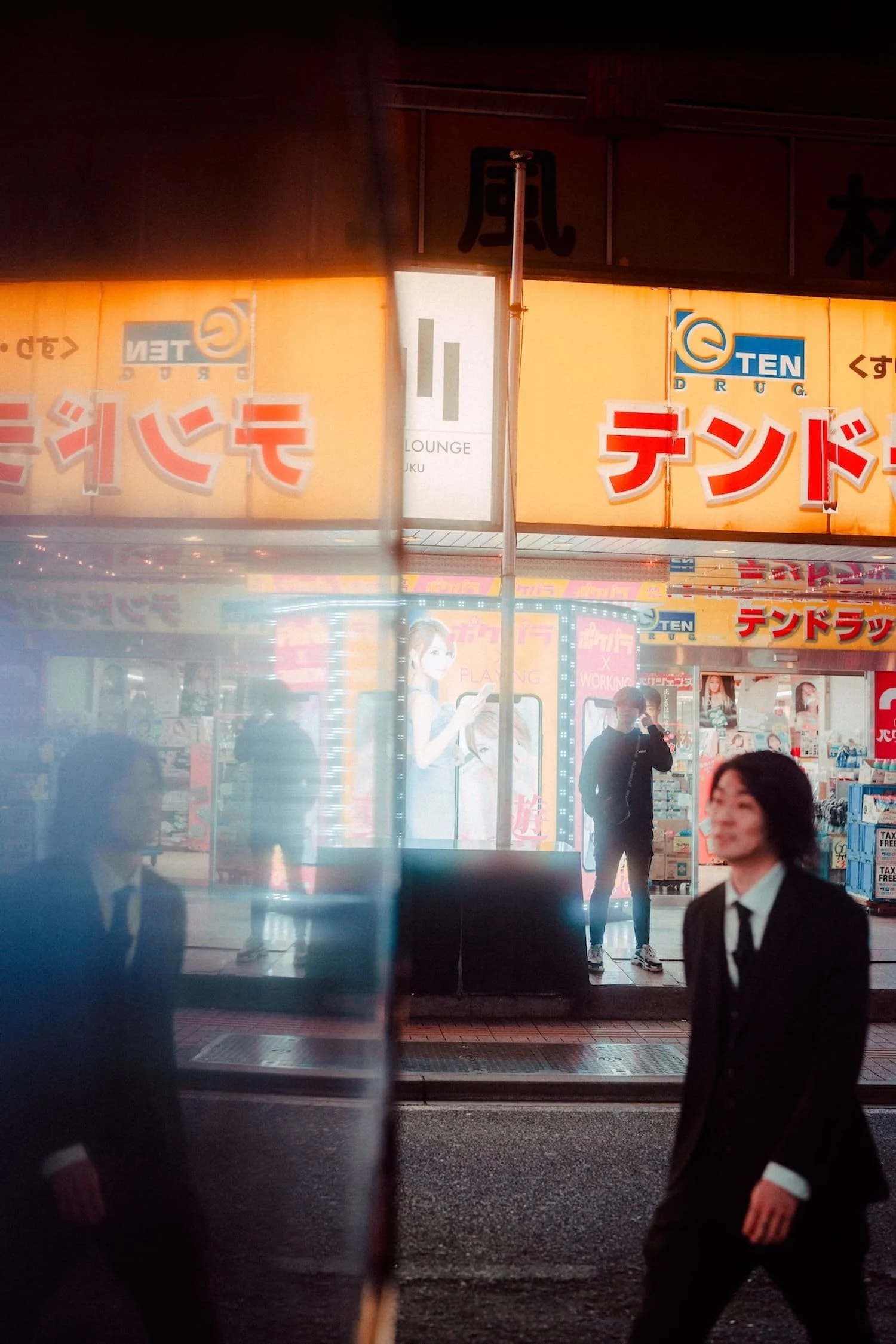 A city street scene at night with a reflection in a glass window showing people walking and standing, illuminated signs in Japanese, and storefronts including a drugstore.
