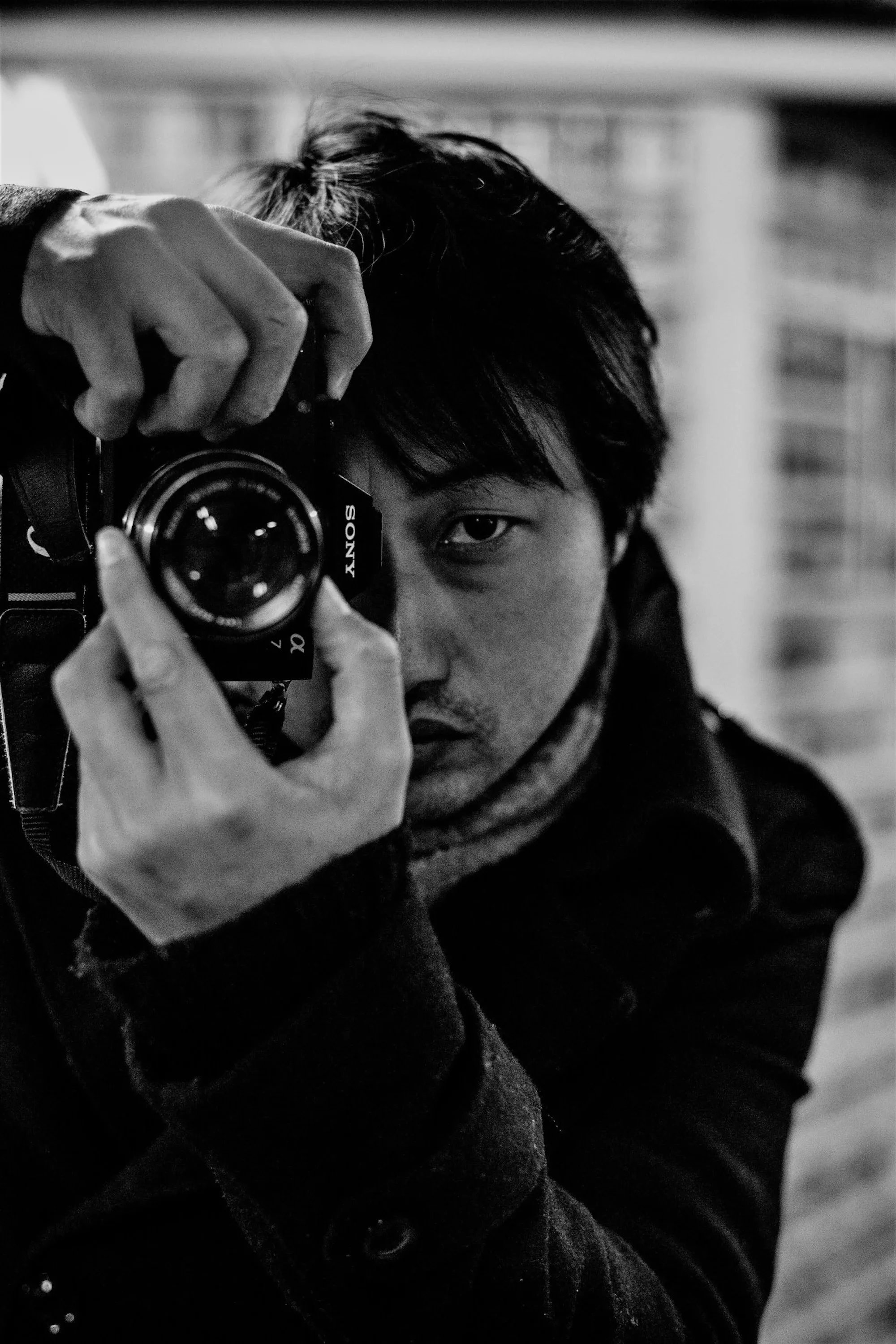 A person with dark hair and an eye visible, taking a mirror selfie with a camera, wearing a dark coat and scarf in an indoor setting.