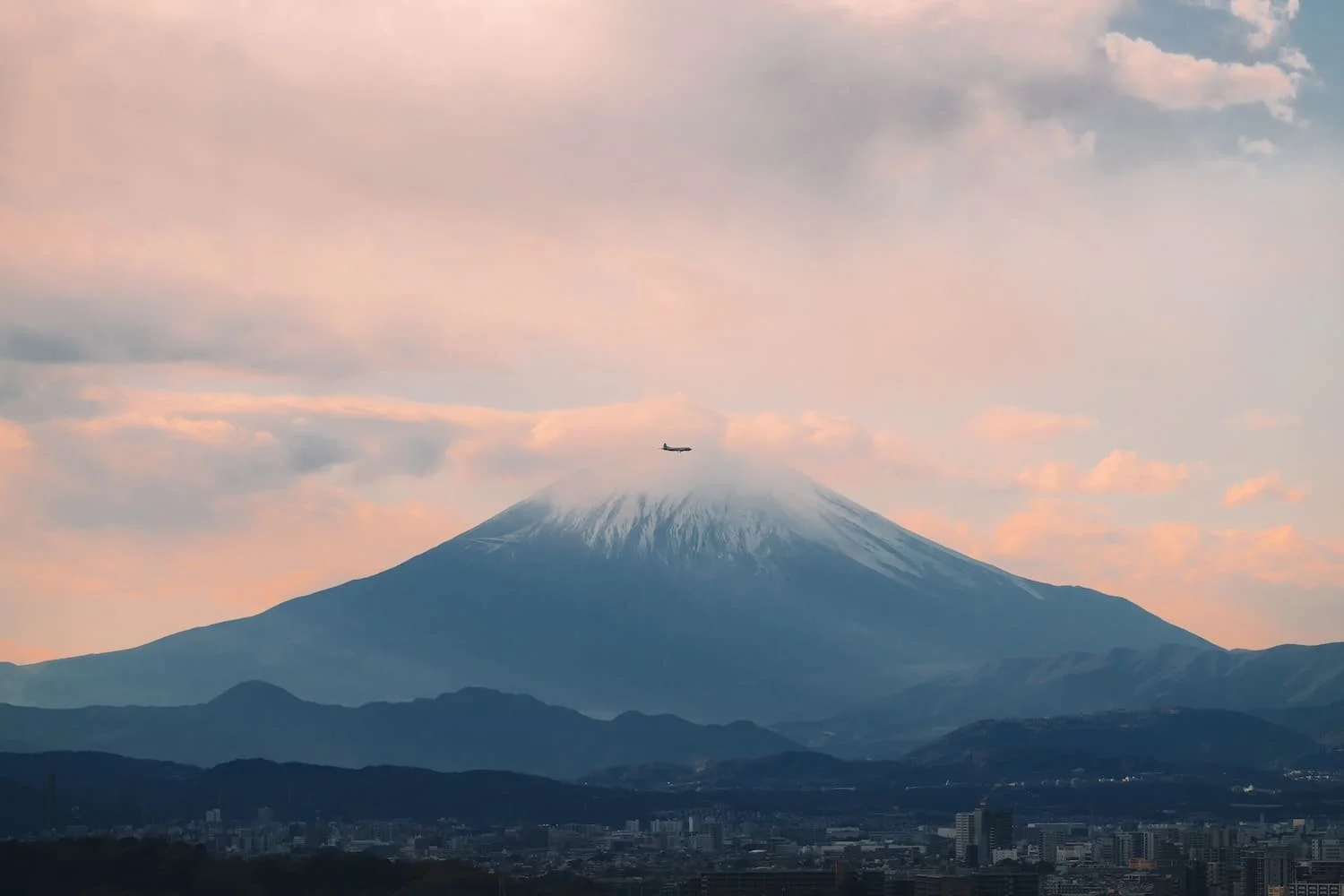 A snow-capped mountain with an airplane flying in front of it, under a cloudy sky.