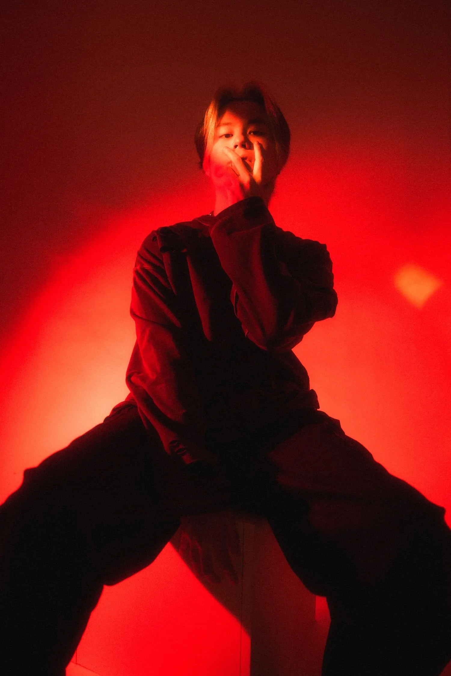 Man sitting on a stool with legs apart, wearing dark clothing, illuminated by red lighting, with hand partially covering face in a low-angle shot by studio photographer Deniz Demir