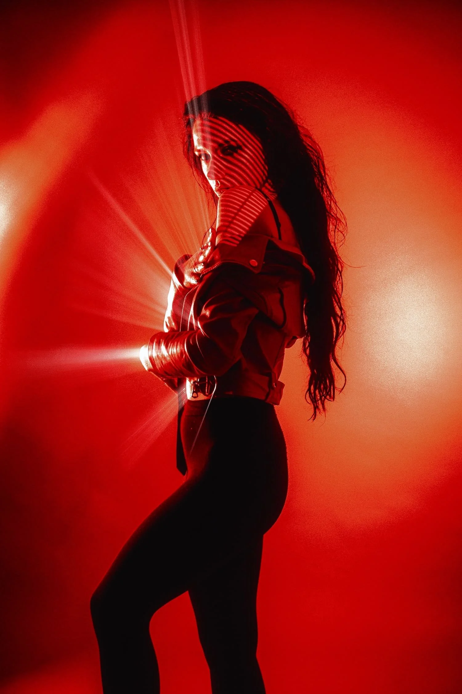 A woman with long dark curly hair stands against a vivid red background, wearing a jacket and tight black pants, with streaks of light and shadow creating a dramatic effect.