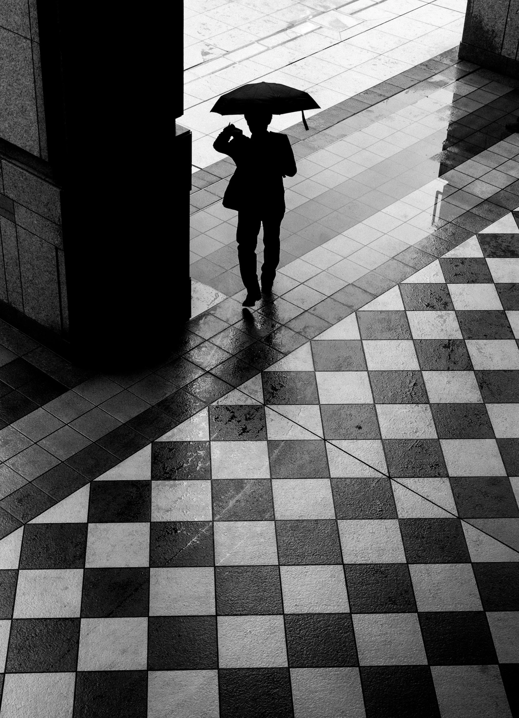 Silhouette of a person holding an umbrella on a rainy day, standing on wet tiled pavement in an urban setting.