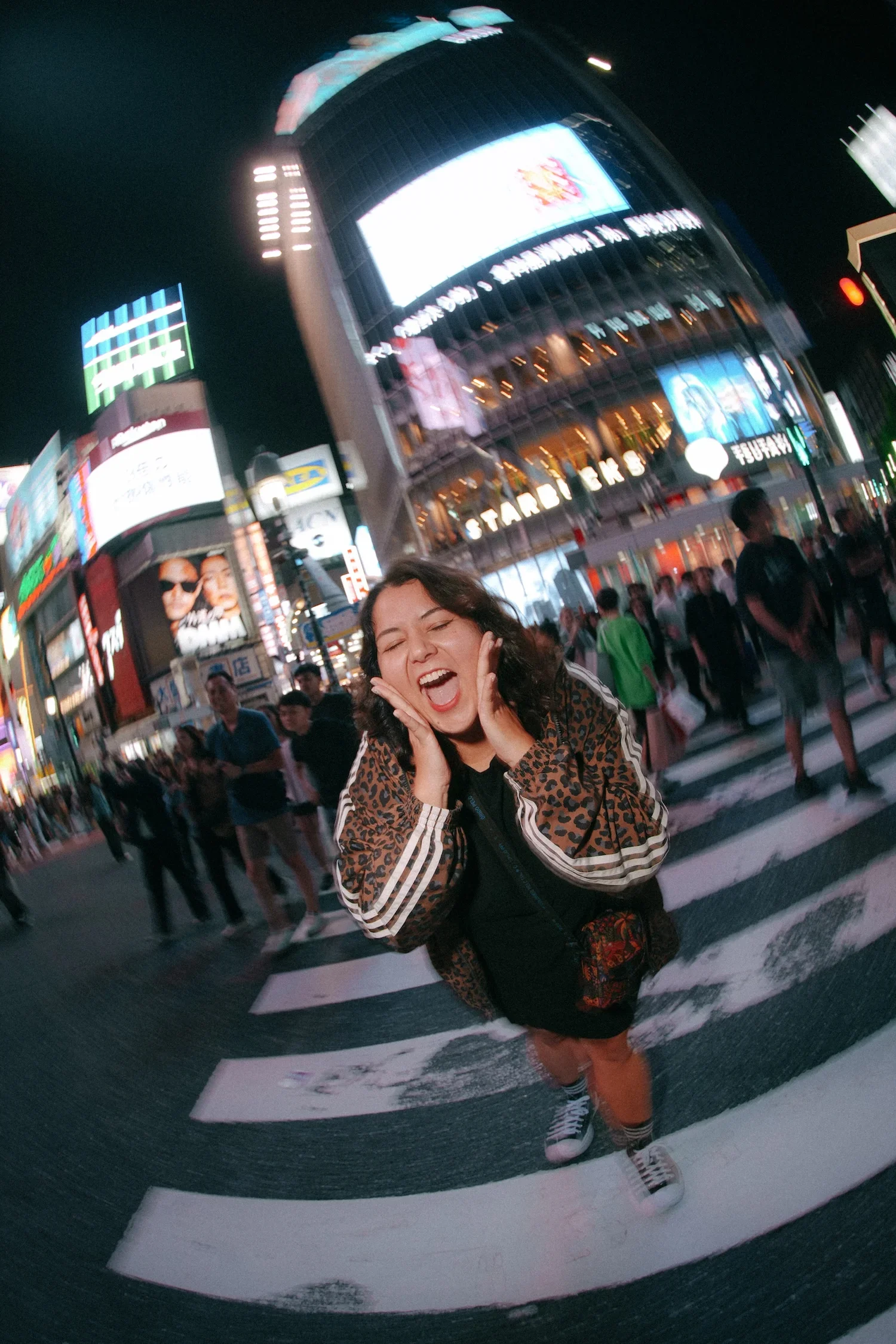 A woman with curly hair smiling with her eyes closed, holding her face with both hands, standing on a city street at night with digital billboards and a crowd of people in the background.