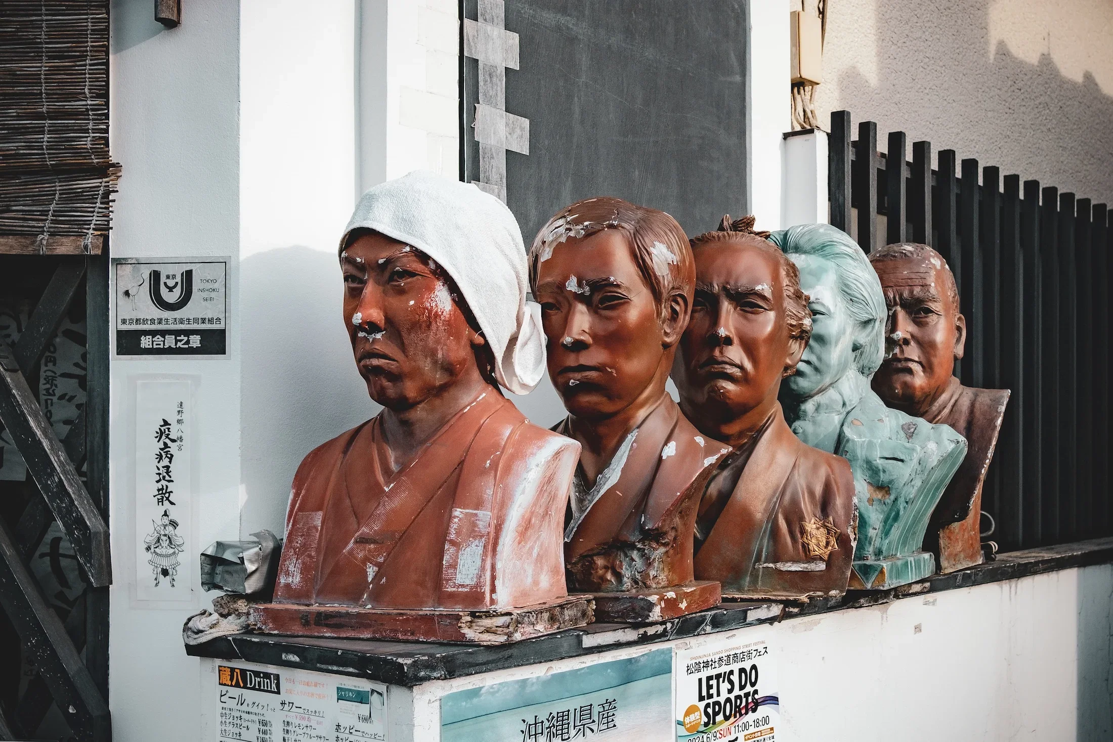 A display of several bust sculptures of people, some with bandages or cloth on their heads, arranged on a shelf outdoors. The background includes a white wall, posters with Japanese writing, and a black fence.